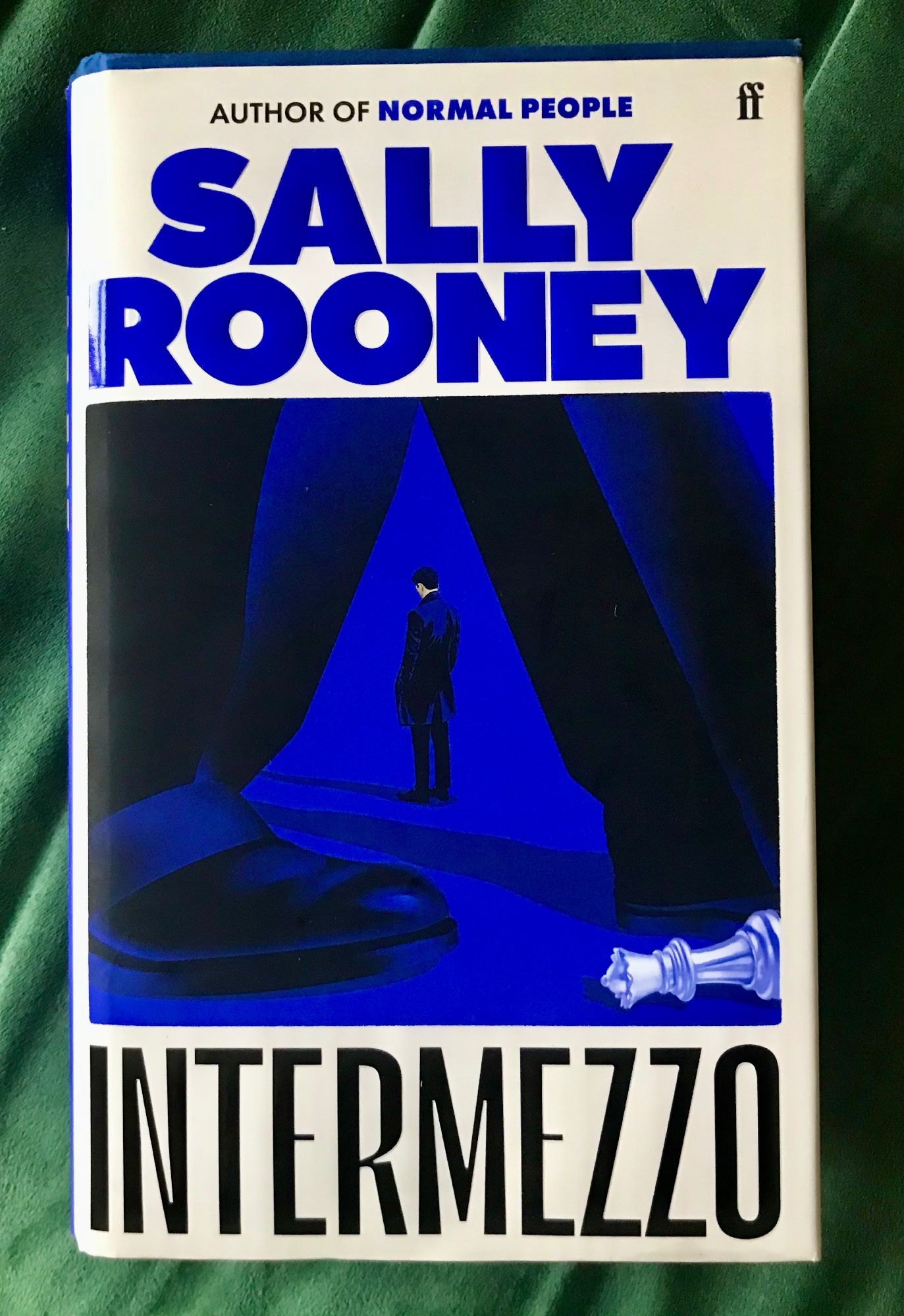 Hardback edition of Intermezzo by Sally Rooney (Faber), with a strange blue-black illustration of a dark-haired white-skinned man in a coat looking sadly downwards, seen between the striding legs of a man in trousers and smart shoes or boots, with a white chess piece (the queen) lying on its side.