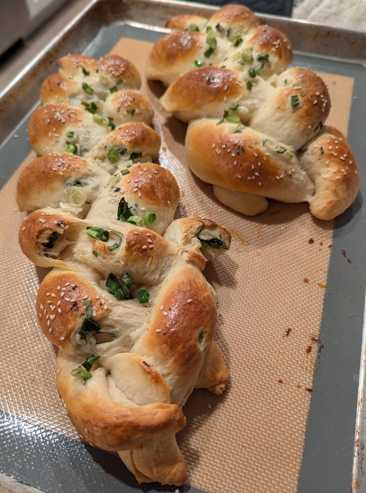 Two loaves of bread shaped as 3-stranded braids, fresh out of the oven. They are sprinkled with sesame seeds and scallions and smell delightful.