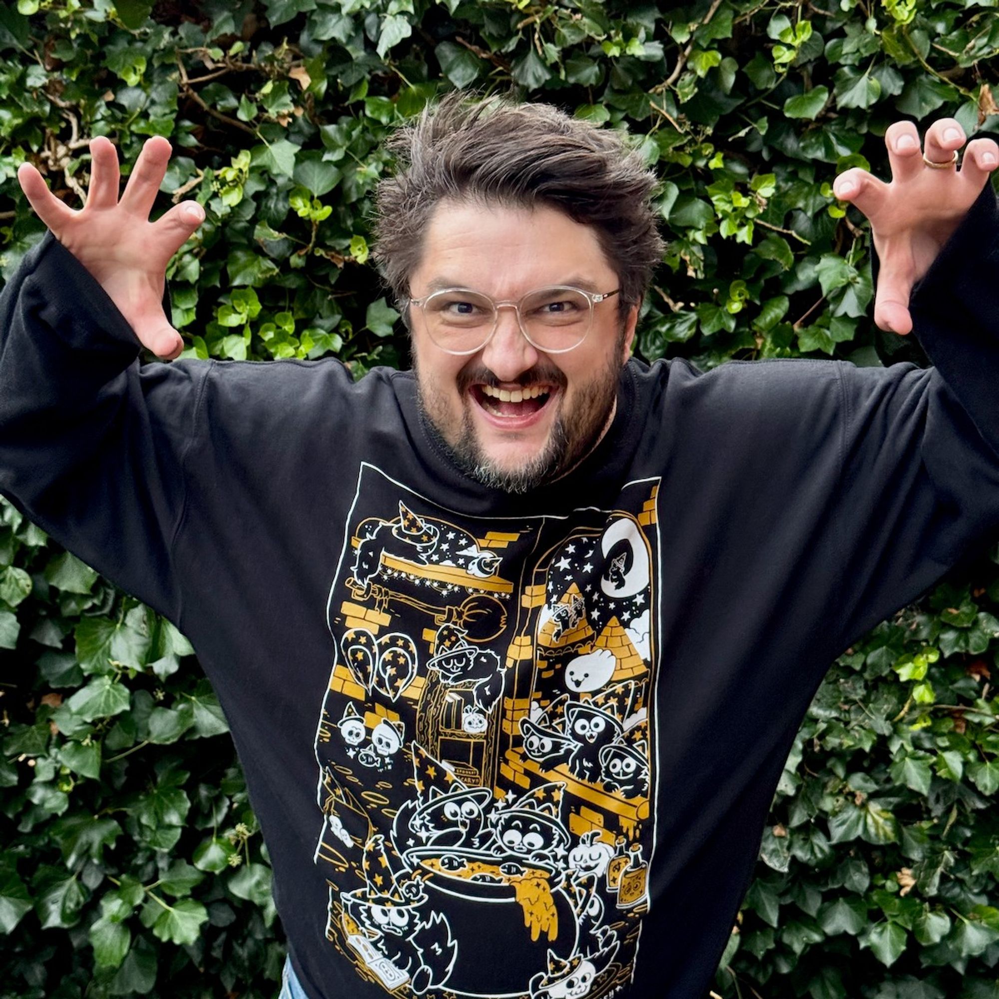 Stef is posing in the wizard cats black sweater, doing spooky claw hands in front of an ivy bush.