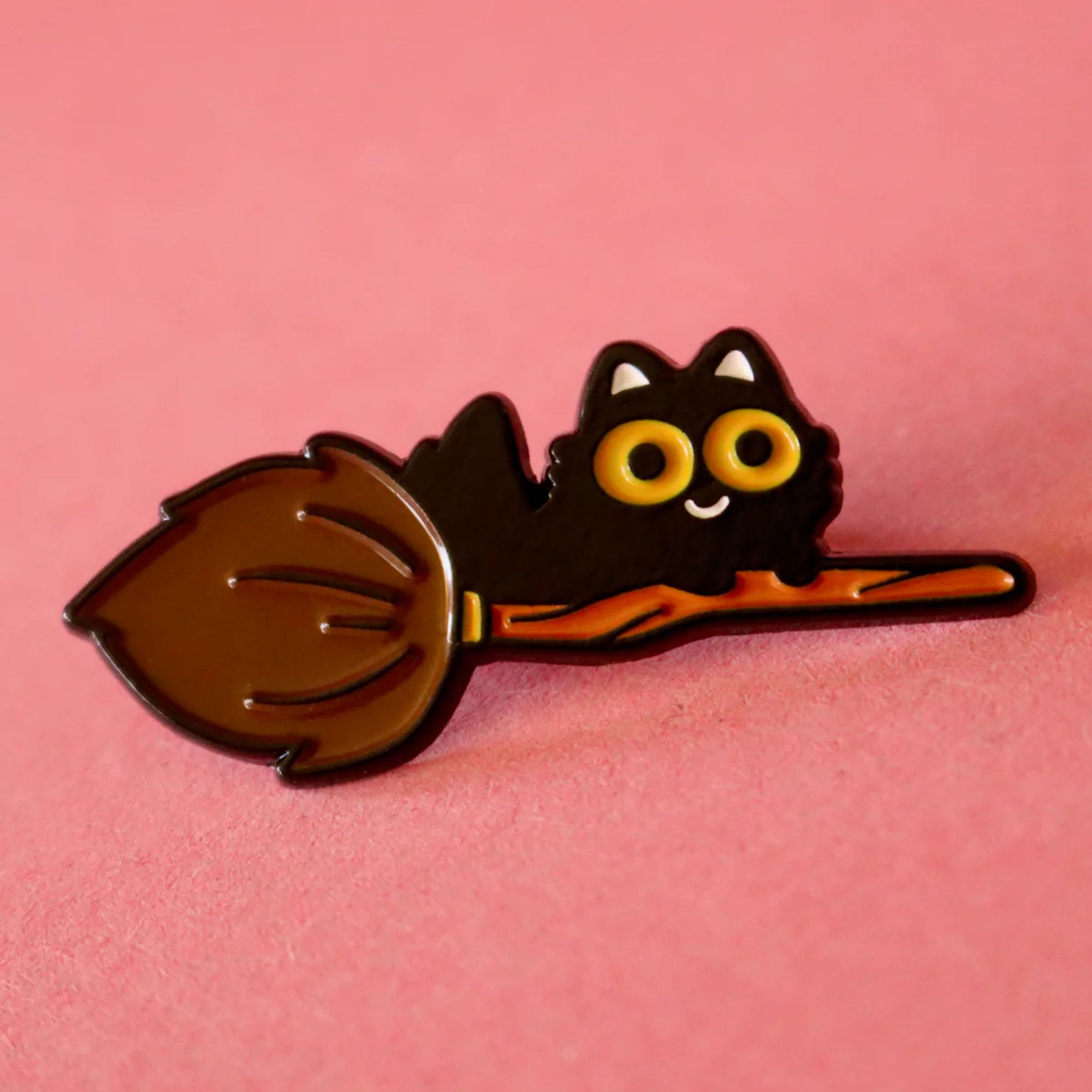 A soft enamel pin with black outlines! A small fluffy cat with big orange eyes is riding on a broomstick.