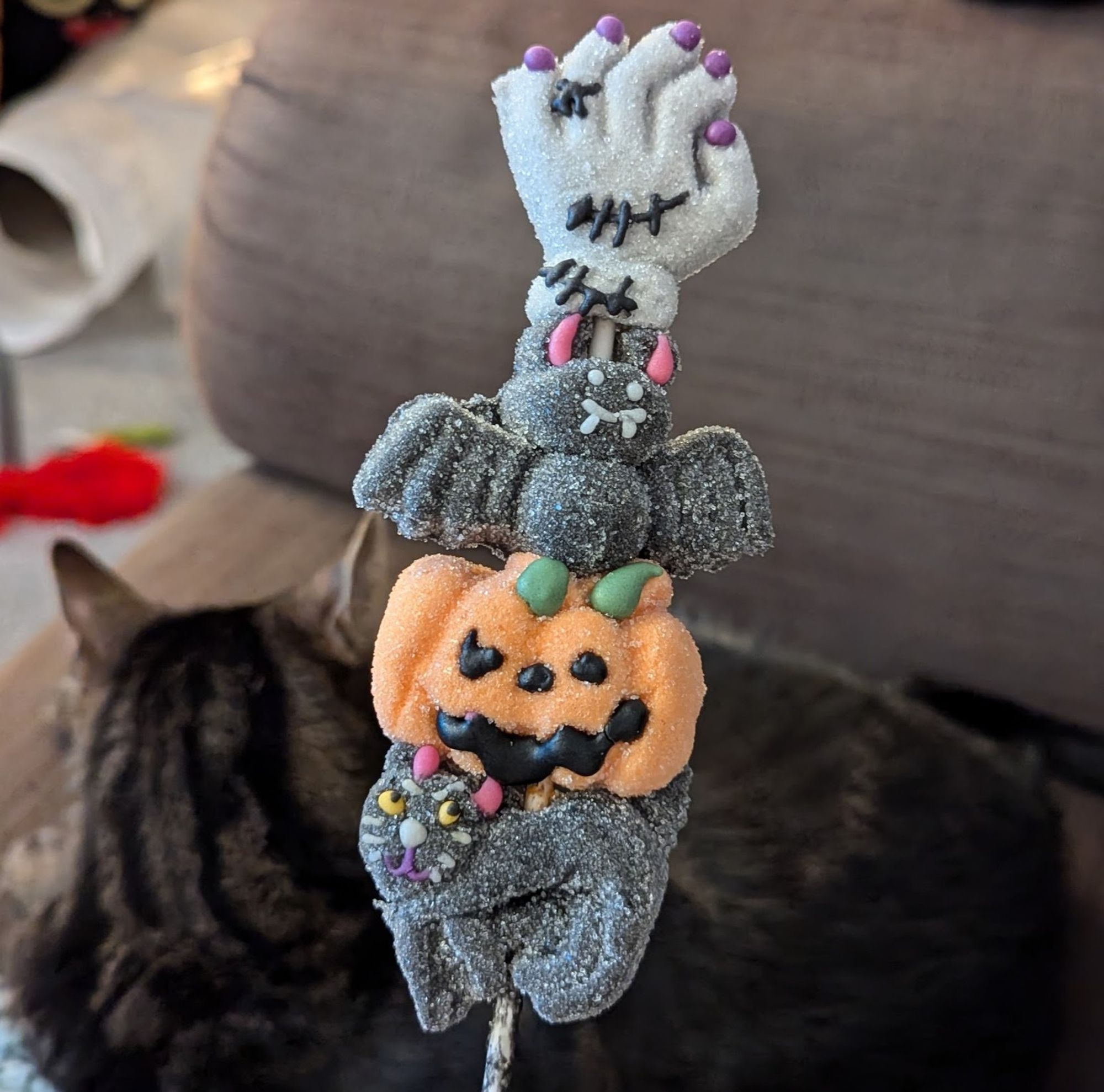 A jelly sugary stick treat thing? It's made up of marshmallows, the top has a zombie hand, followed by a bat, followed by a pumpkin, ending with a black cat. Pesto the cat is blurry in the background looking away from the camera.