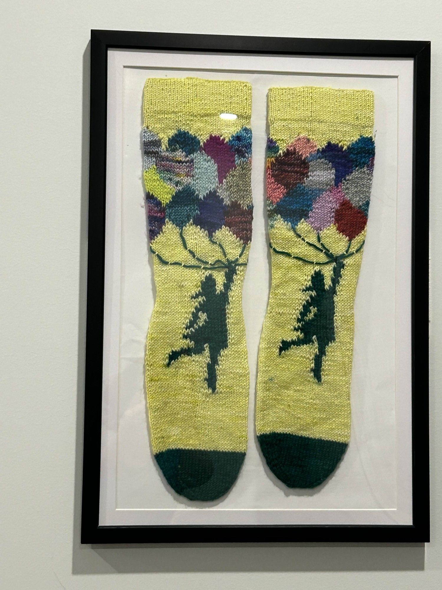 A pair of hand knit socks, each sock showing a pattern of a girl holding a large bunch of multicolored balloons, hung on the wall inside a picture frame