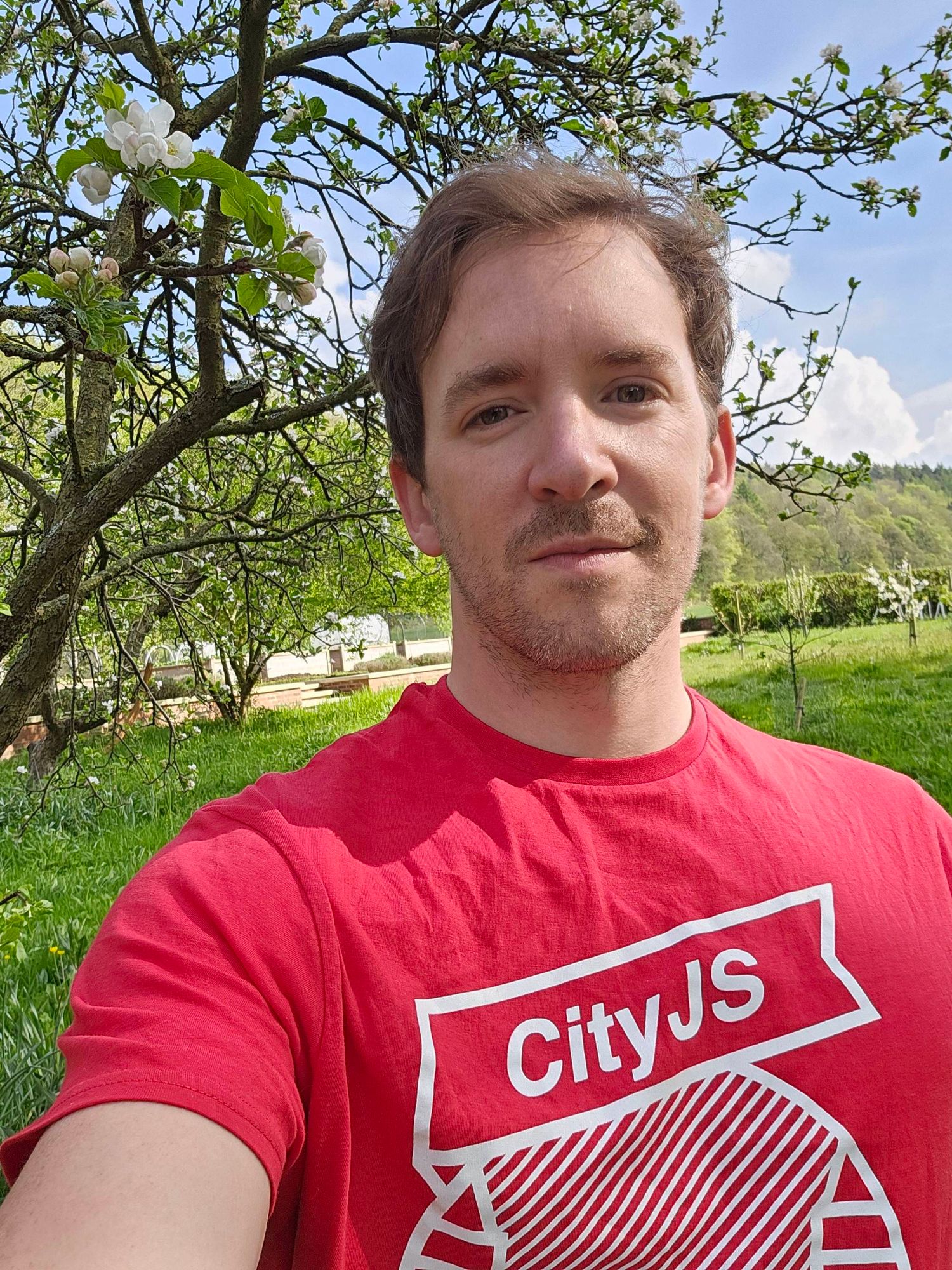 A selfie of me under an apple tree, with blue sky and trees in the background. I'm wearing a red CityJS T-shirt.