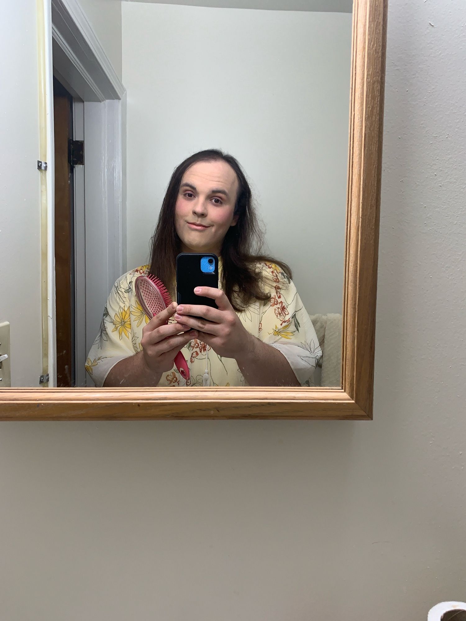 A picture of Cecily from spring 2022, not long before beginning hormones, in her friends bathroom getting ready for a performance with her band. She has long brown hair (which has been a constant for her since the age of 15) and is wearing a sheer yellow floral top with a short sleeve button down underneath it. She has just done her make up for the first time since having lived with the person whose mirror this was more than a year prior and will get better at it