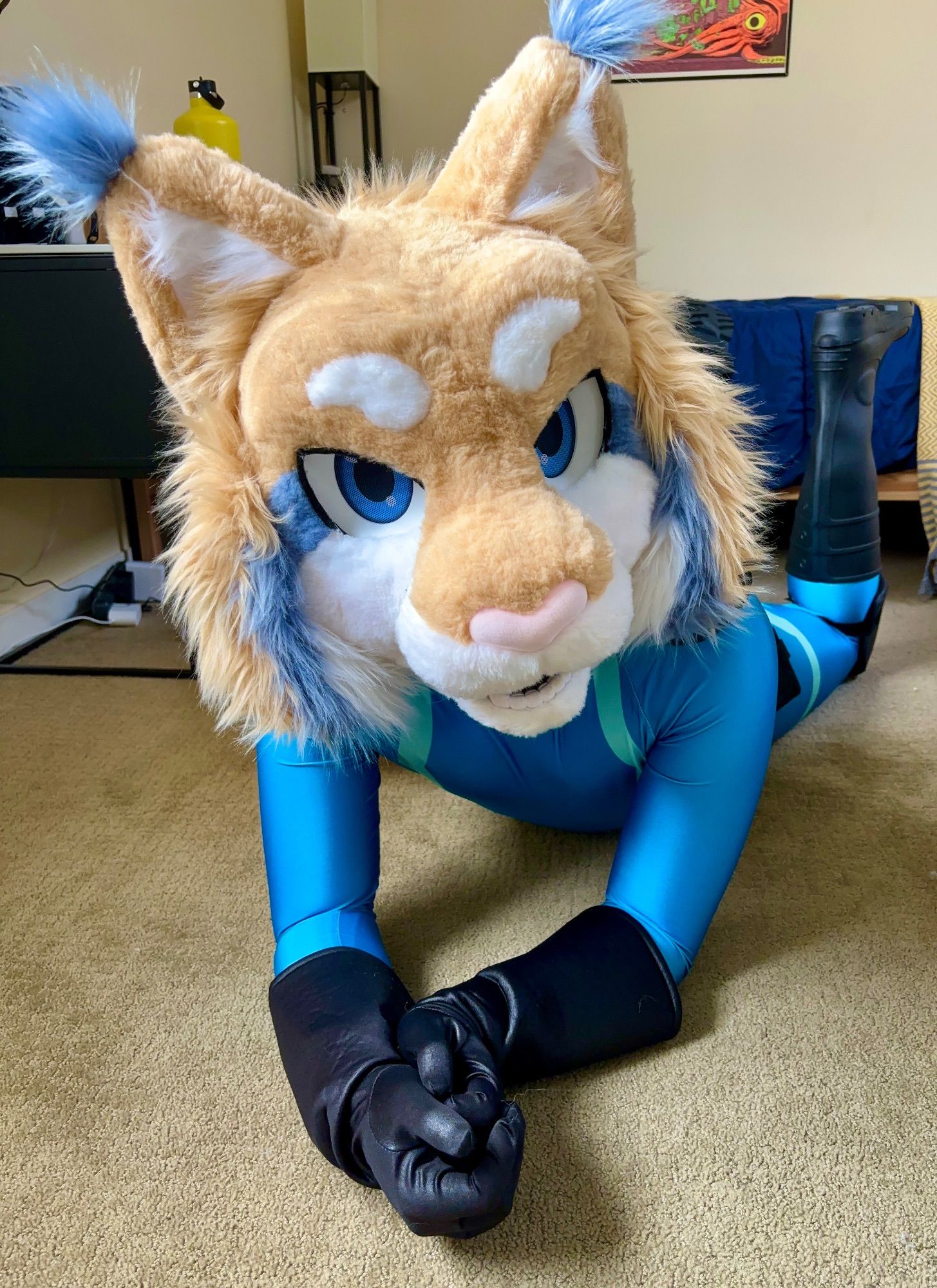 A tan feline lynx fursuiter wearing a blue spandex superhero suit with black boots and gloves lies on his stomach, turned up towards the camera and his boots pointing upwards.