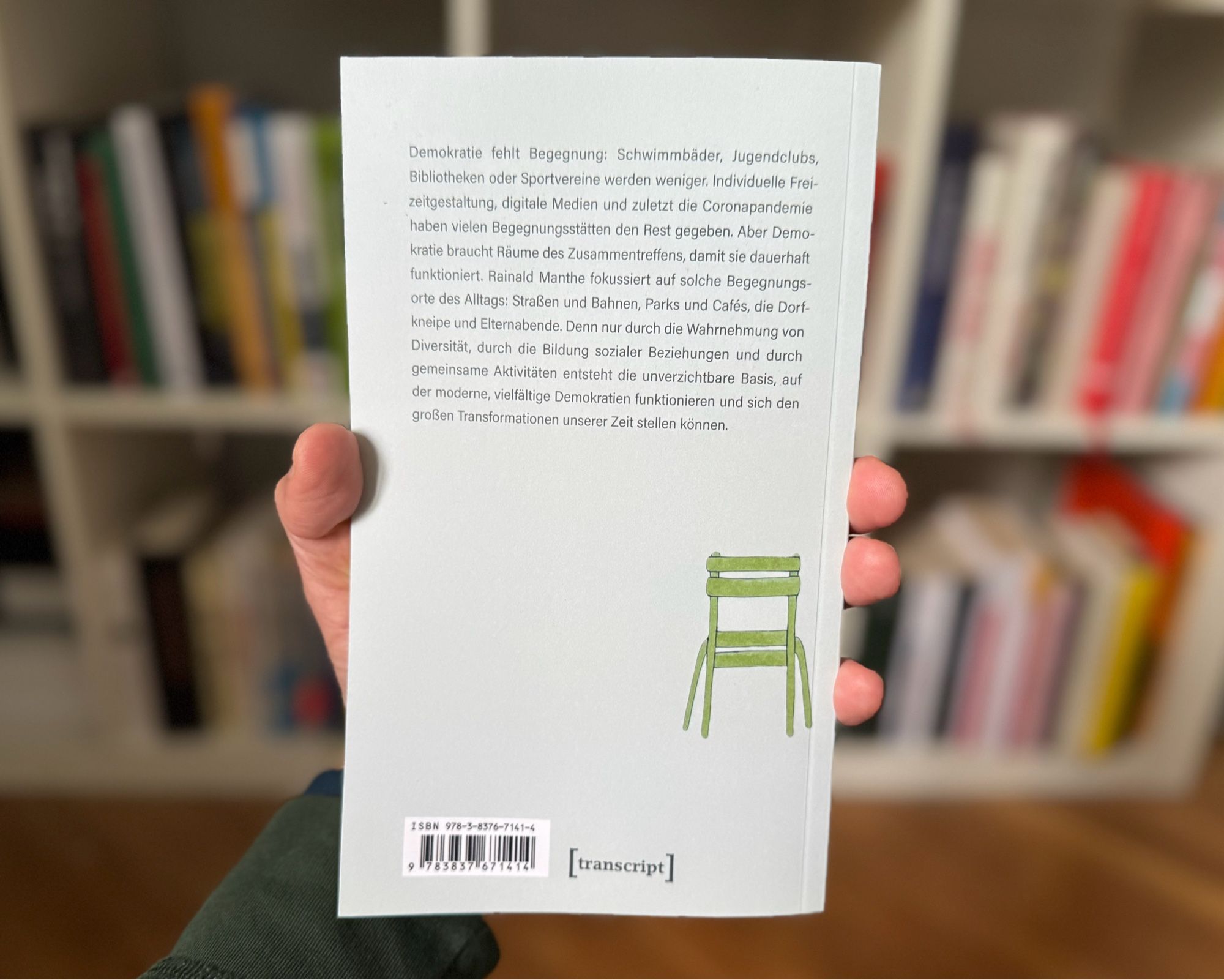 Das Bild zeigt die Rückseite des Buches „Demokratie fehlt Begegnung“ von Rainald Manthe, das von meiner Hand vor einem Bücherregal gehalten wird. Auf der Rückseite befindet sich folgender Text: „Demokratie fehlt Begegnung: Schwimmbäder, Jugendclubs, Bibliotheken oder Sportvereine werden weniger. Individuelle Frei-zeitgestaltung, digitale Medien und zuletzt die Coronapandemie haben vielen Begegnungsstätten den Rest gegeben. Aber Demokratie braucht Räume des Zusammentreffens, damit sie dauerhaft funktioniert. Rainald Manthe fokussiert auf solche Begegnungs-orte des Alltags: Straßen und Bahnen, Parks und Cafés, die Dorf-kneipe und Elternabende. Denn nur durch die Wahrnehmung von Diversität, durch die Bildung sozialer Beziehungen und durch gemeinsame Aktivitäten entsteht die unverzichtbare Basis, auf der moderne, vielfältige Demokratien funktionieren und sich den großen Transformationen unserer Zeit stellen können.“
Darunter ist ein Barcode mit der ISBN-Nummer (978-3-8376-7141-4) zu sehen.