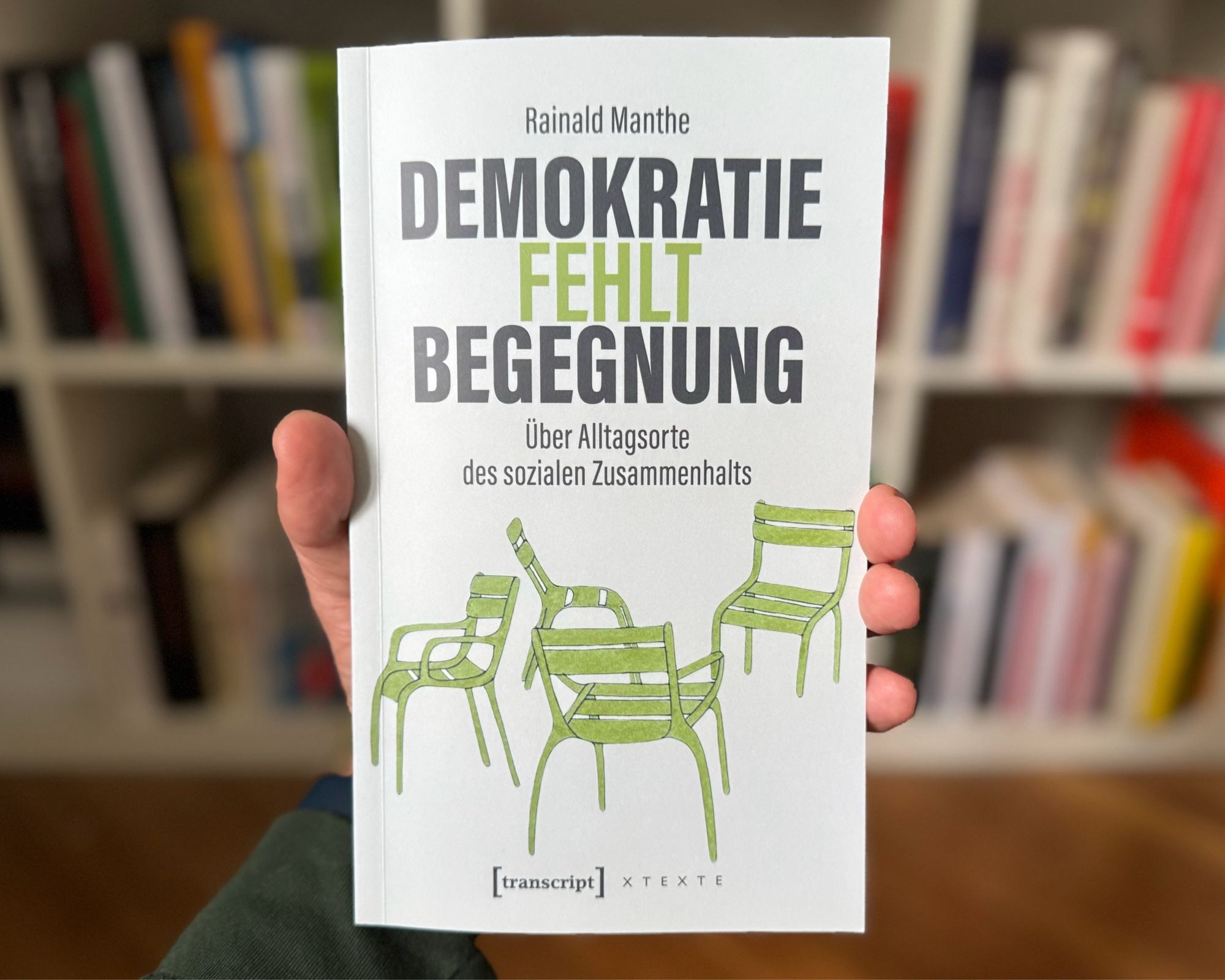 Das Bild zeigt meine Hand, die ein Buch vor einem Bücherregal hält. Der Titel des Buches lautet „Demokratie fehlt Begegnung: Über Alltagsorte des sozialen Zusammenhalts“ von Rainald Manthe. Auf dem Buchcover sind grüne Illustrationen von vier Stühlen abgebildet, die in unterschiedlichen Positionen angeordnet sind. Der Titel ist in zwei Schriftfarben gestaltet: „Demokratie“ und „Begegnung“ sind schwarz, während „fehlt“ in grün hervorgehoben ist. Der Untertitel befindet sich unterhalb des Haupttitels und beschreibt das Thema des Buches, das sich mit Orten beschäftigt, an denen sozialer Zusammenhalt im Alltag stattfindet. Am unteren Rand des Buchcovers ist das Logo des Verlags „[transcript]“ sichtbar. Im Hintergrund des Bildes sind unscharf weitere Bücher in einem Regal zu erkennen.