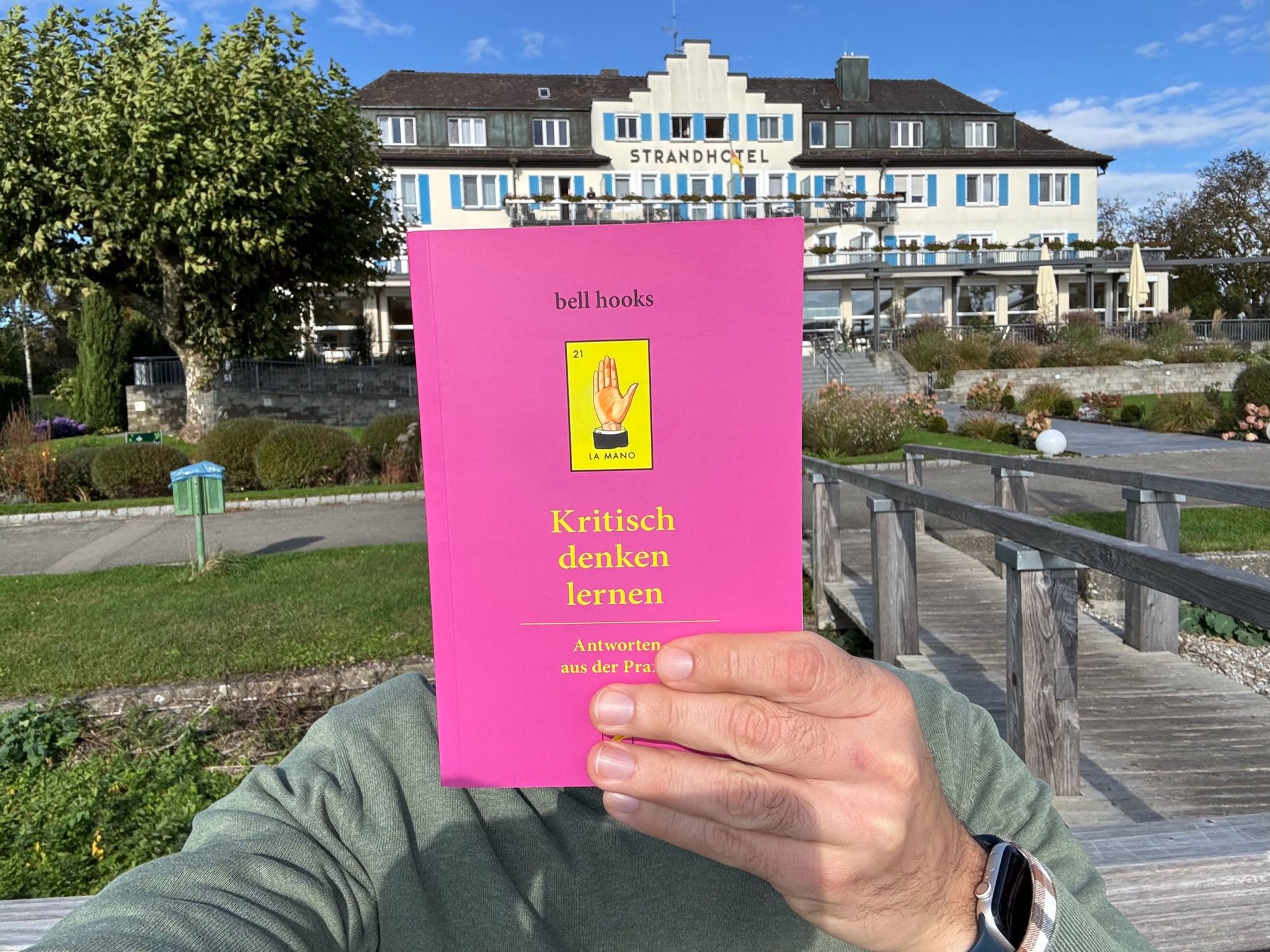 Das Bild zeigt mich, vor einem großen, eleganten Gebäude, auf dem „Strandhotel“ steht. Ich halte ein Buch hoch, das den Titel „Kritisch denken lernen“ von bell hooks trägt. Das Buch ist leuchtend pink und verdeckt mein Gesicht, sodass nur die Hand und der Arm sichtbar sind. Im Hintergrund sieht man eine Promenade und gepflegte Gärten.