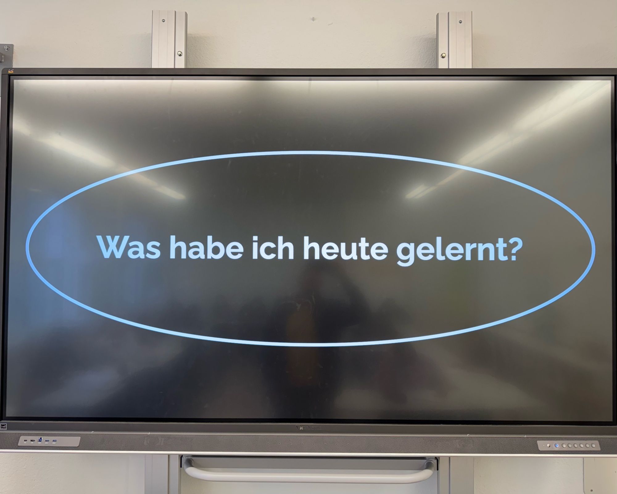 Das Bild zeigt einen großen Bildschirm, auf dem eine Frage in der Mitte dargestellt wird: "Was habe ich heute gelernt?" Die Frage ist in weißer Schrift auf einem schwarzen Hintergrund geschrieben, umgeben von einer ovalen, blauen Umrandung. Der Bildschirm scheint ein interaktives Whiteboard oder ein digitales Präsentationsgerät zu sein, das in einem Klassenzimmer oder Schulungsraum verwendet wird. Oberhalb des Bildschirms sind Halterungen oder Befestigungen zu erkennen, die den Bildschirm an der Wand befestigen. Der Raum spiegelt sich leicht im Bildschirm, aber es sind keine Personen direkt sichtbar.