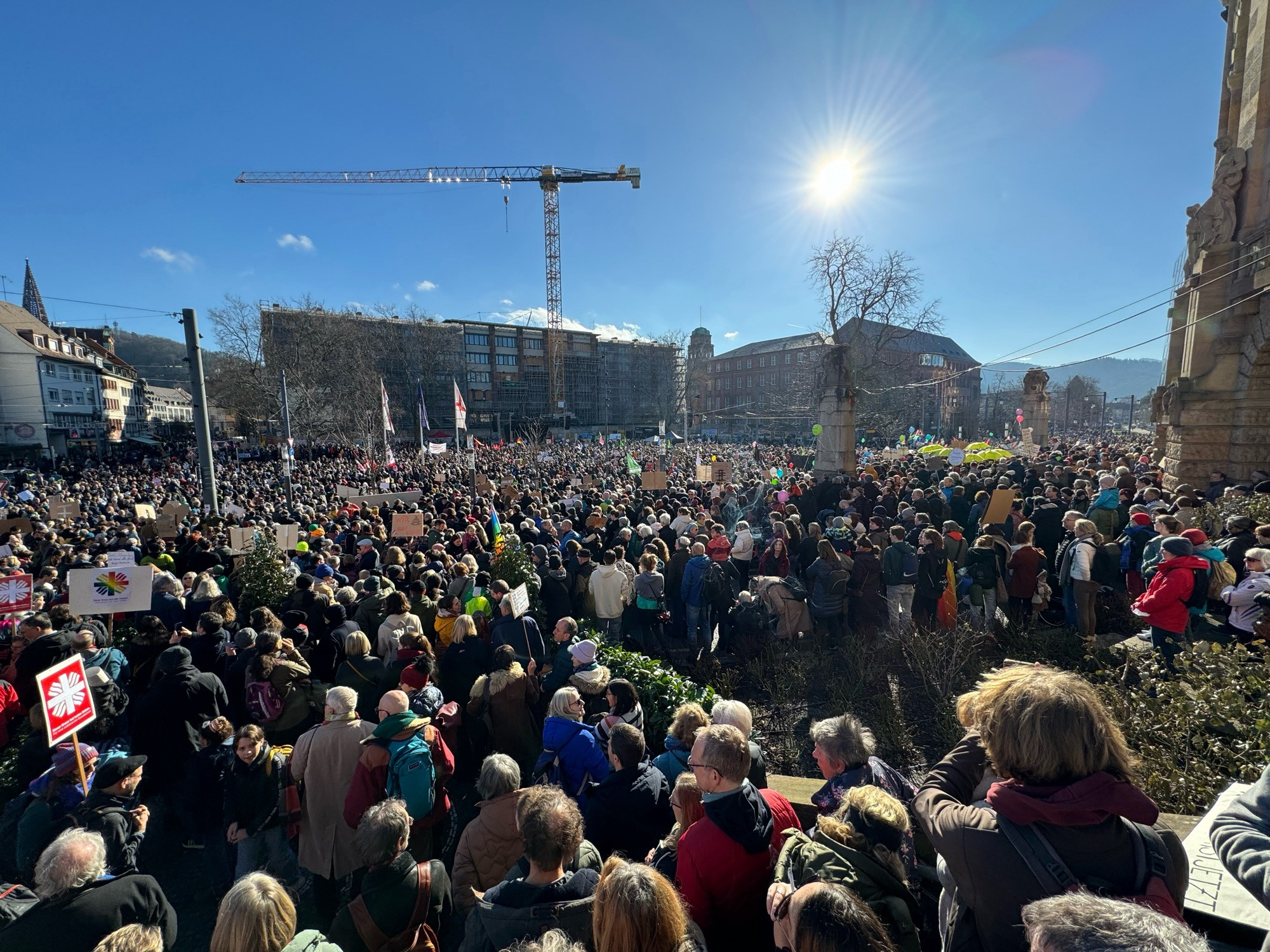 Das Bild zeigt eine Menschenmenge bei einer Demonstration oder öffentlichen Versammlung an einem sonnigen Tag. Viele Teilnehmer halten Plakate und Flaggen, wobei einige Symbole wie die Friedenstaube und die Sonnenblume zu erkennen sind, die oft für Frieden oder ökologische Bewegungen stehen. Im Hintergrund sind städtische Gebäude und ein Baukran sichtbar, was darauf hindeutet, dass das Ereignis in einem städtischen Umfeld stattfindet. Die Sonne scheint hell am klaren Himmel, und es gibt keine Anzeichen für Gewalt oder Unruhen, was auf eine friedliche Veranstaltung hindeutet.