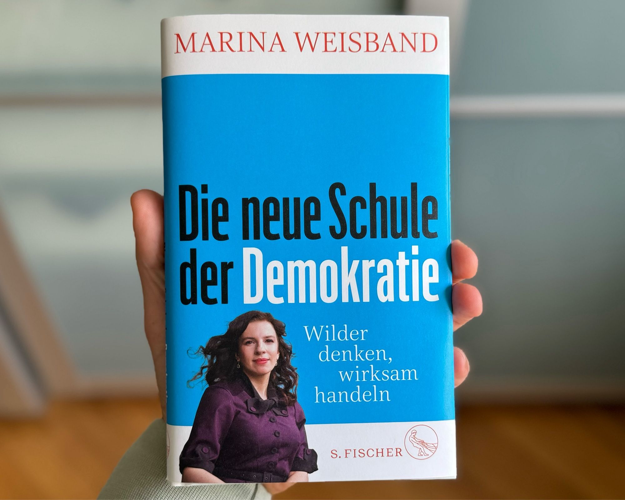 Das Bild zeigt meine Hand, die ein Buch hält. Das Buch hat einen blauen Umschlag mit weißer und roter Schrift. Der Titel des Buches lautet "Die neue Schule der Demokratie" und der Autorenname "Marina Weisband" steht in großen roten Buchstaben darüber. Unter dem Titel steht der Untertitel "Wilder denken, wirksam handeln" in weißer Schrift. Auf dem Umschlag ist ein Foto von Marina Weisband zu sehen, die eine violette Bluse trägt und lächelt. Im unteren Bereich des Umschlags befindet sich das Logo des S. Fischer Verlags. Der Hintergrund hinter dem Buch ist unscharf und lässt keine weiteren Details erkennen.