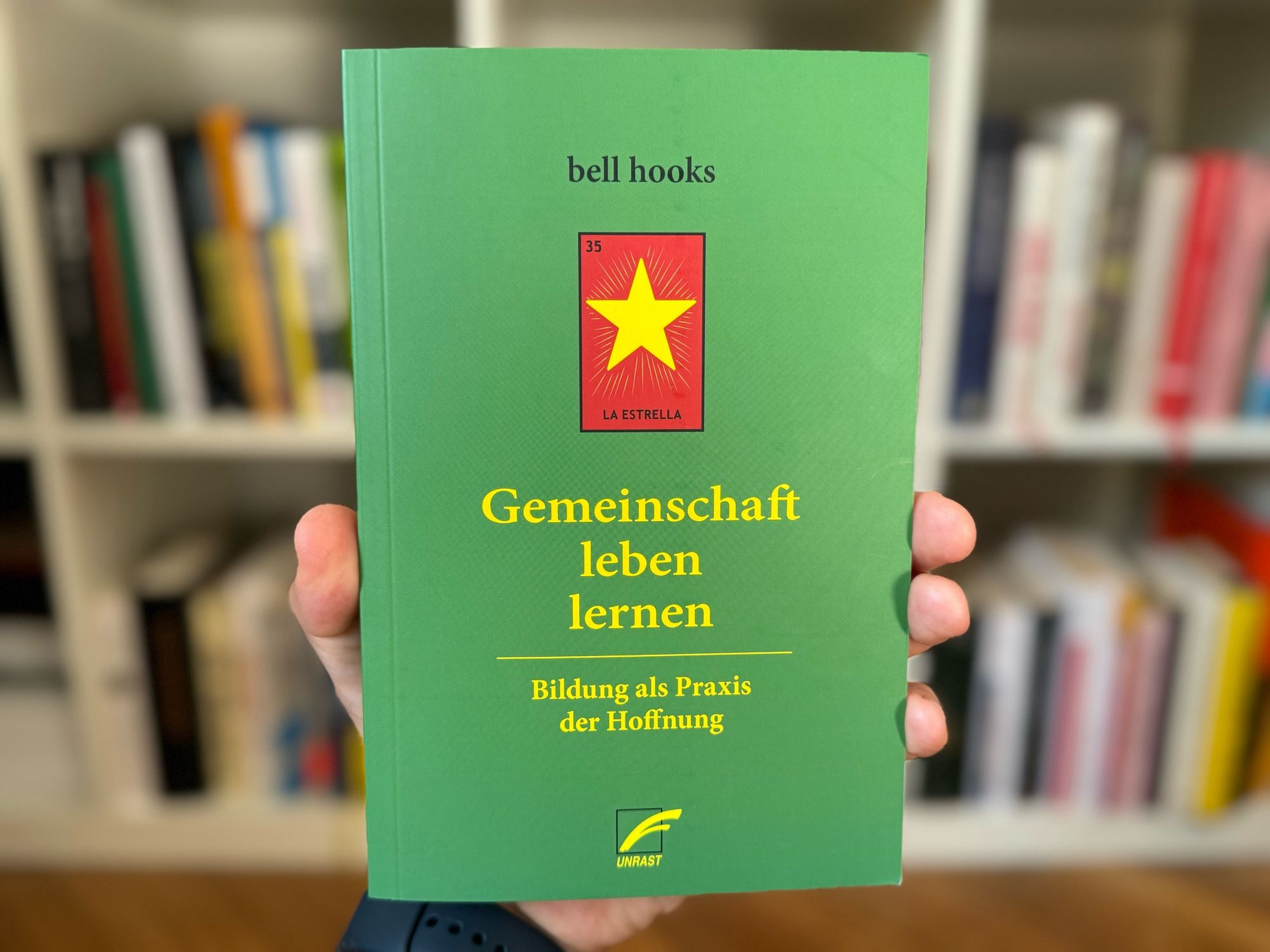 Man sieht ein Foto einer Vorderseite eines Buches: Das Buch hat einen grünen Einband. Der Titel des Buches lautet „Gemeinschaft leben lernen“. Untertitel: „Bildung als Praxis der Hoffnung“. Die Autorin ist bell hooks. Auf dem Cover befindet sich ein rotes Quadrat mit einem gelben Stern in der Mitte und dem Text „LA ESTRELLA“ in der oberen linken Ecke. Die Nummer „35“ steht in einer kleinen schwarzen Box über dem Stern. Unten rechts auf dem Cover befindet sich das Logo des Verlages „Unrast“.