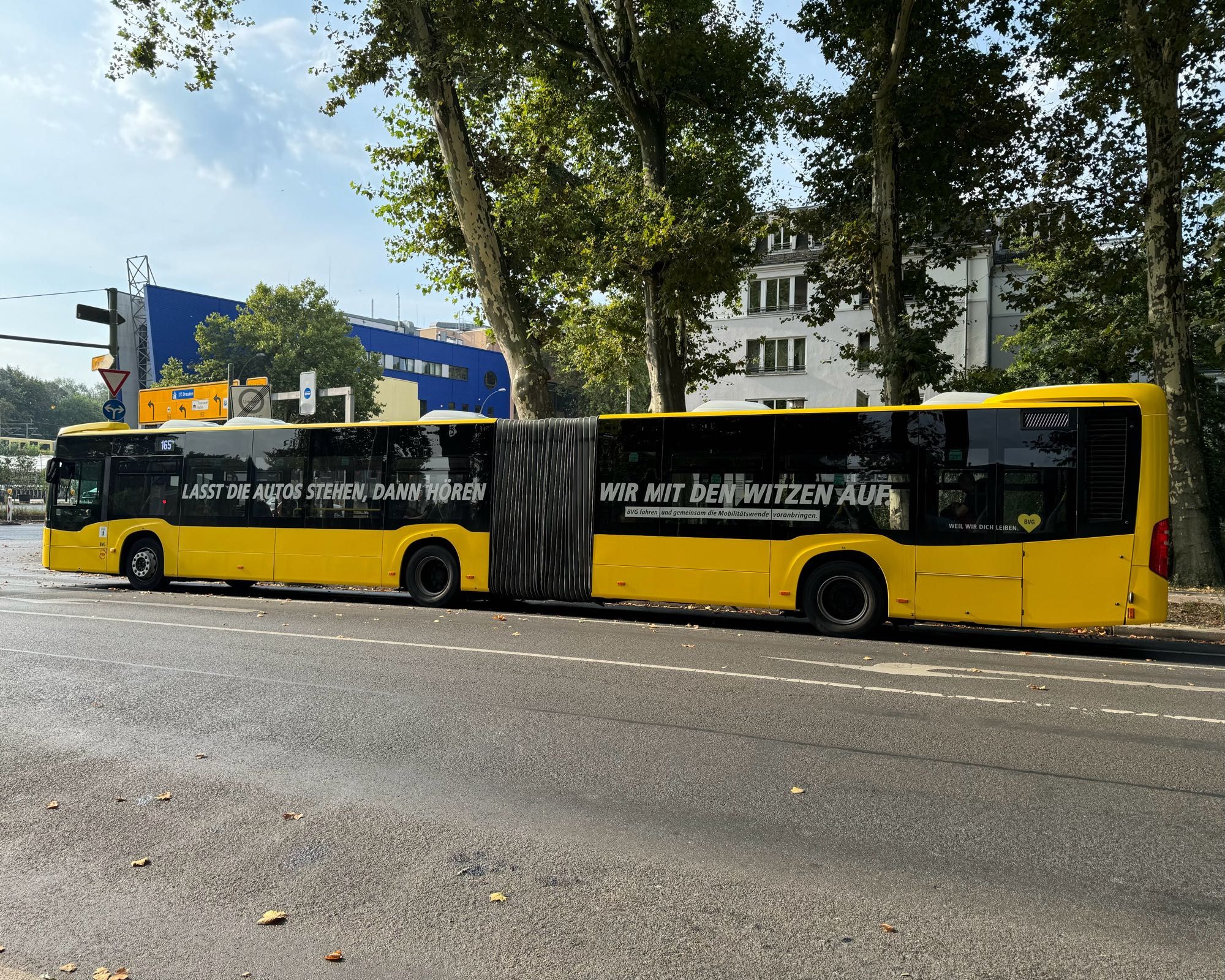 Das Bild zeigt einen langen, gelben Gelenkbus, der am Straßenrand geparkt ist. Der Bus hat eine auffällige schwarze Werbebotschaft entlang der Seite. Der Text lautet: "LASST DIE AUTOS STEHEN, DANN HÖREN WIR MIT DEN WITZEN AUF." Darunter befinden sich kleinere Textzeilen, die in kleinerer Schrift geschrieben sind, aber diese sind aufgrund der Bildqualität schwer lesbar. Der Bus steht auf einer asphaltierten Straße, die von großen Bäumen gesäumt ist. Im Hintergrund sind moderne Gebäude zu sehen, darunter ein blau-weißes Gebäude auf der linken Seite. Das Wetter scheint angenehm zu sein, da die Bäume noch Laub tragen und der Himmel leicht bewölkt ist.