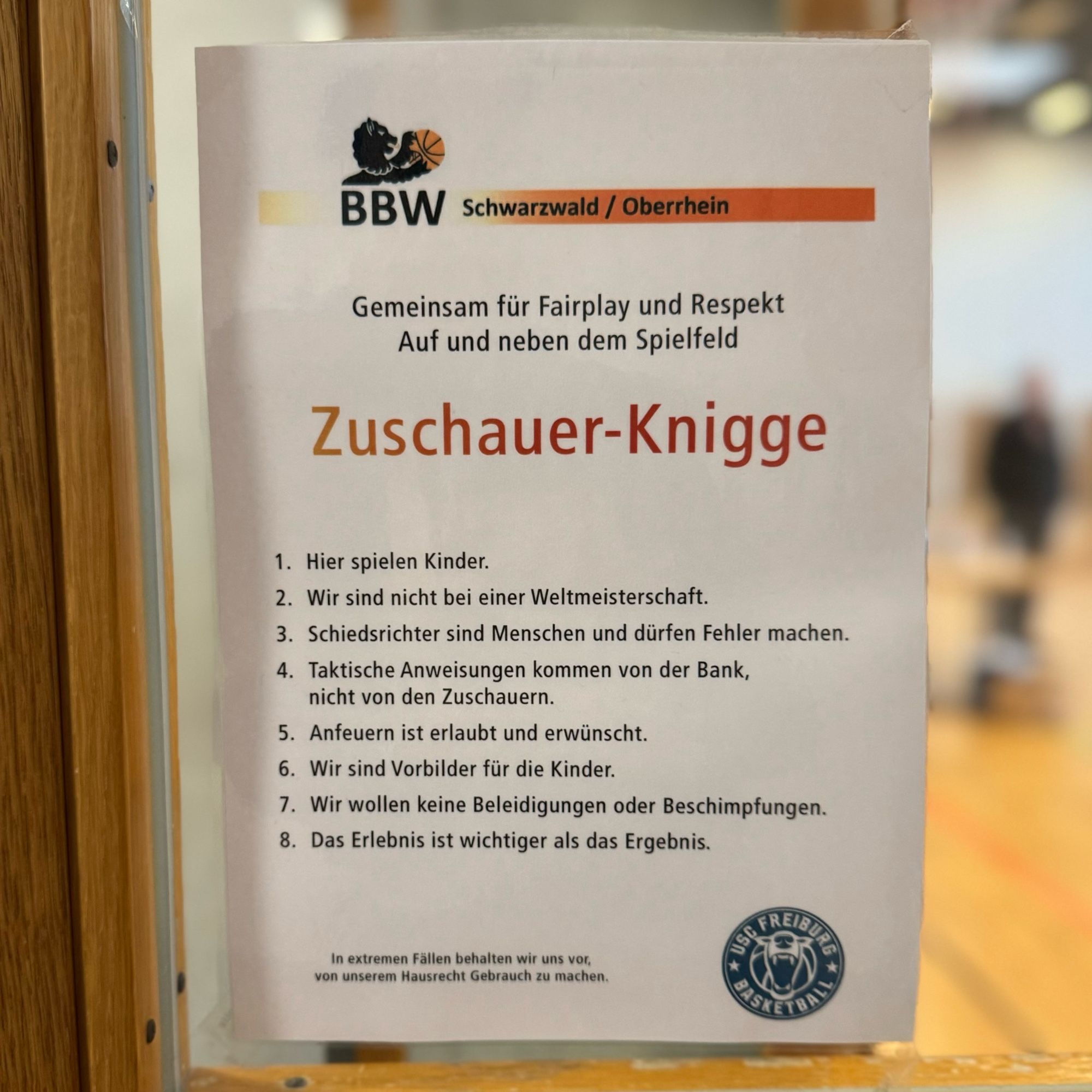 Das Bild zeigt ein Plakat mit dem Titel „Zuschauer-Knigge“, welches im Kontext des Basketballvereins BBW Schwarzwald/Oberrhein angebracht ist. Es richtet sich an Zuschauer und appelliert an Fairplay und Respekt, sowohl auf als auch neben dem Spielfeld. 

Darunter sind acht Regeln aufgelistet:
1. Hier spielen Kinder.
2. Wir sind nicht bei einer Weltmeisterschaft.
3. Schiedsrichter sind Menschen und dürfen Fehler machen.
4. Taktische Anweisungen kommen von der Bank, nicht von den Zuschauern.
5. Anfeuern ist erlaubt und erwünscht.
6. Wir sind Vorbilder für die Kinder.
7. Wir wollen keine Beleidigungen oder Beschimpfungen.
8. Das Erlebnis ist wichtiger als das Ergebnis.

Am unteren Rand des Plakats befindet sich der Hinweis: „In extremen Fällen behalten wir uns vor, von unserem Hausrecht Gebrauch zu machen.“ Rechts unten ist das Logo von USC Freiburg Basketball zu sehen.