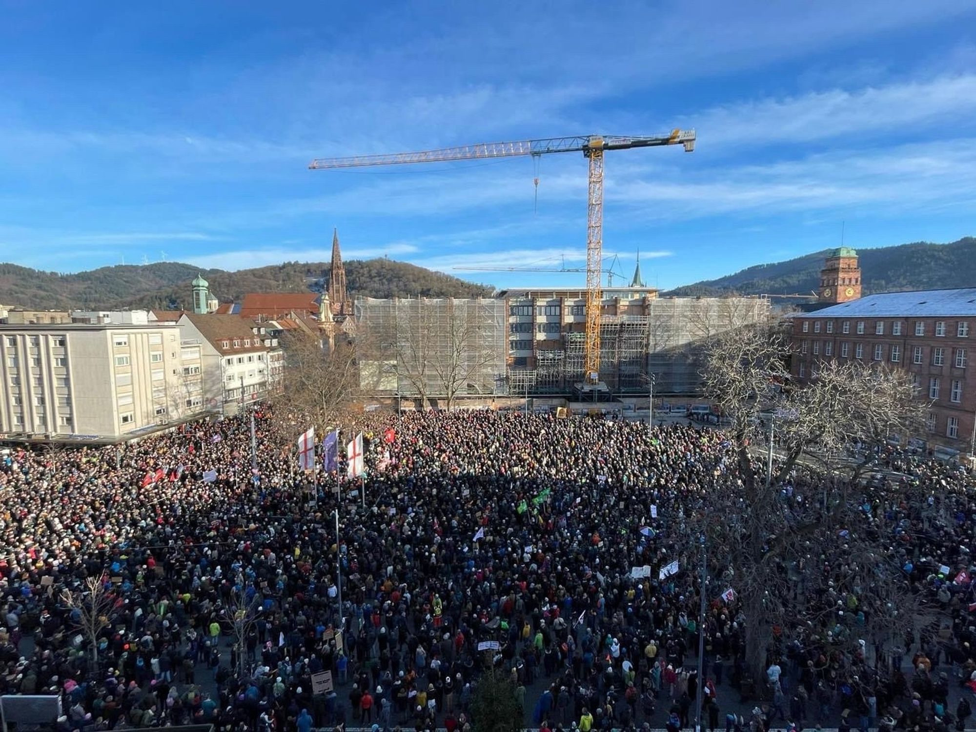 Das Bild zeigt eine große Menschenmenge, die sich in Freiburg rund um den Platz der Alten Synagoge versammelt hat. Im Hintergrund ist eine hügelige Landschaft mit Bäumen und ein klarer blauer Himmel zu sehen. Auf der linken Seite ragt ein spitzer Kirchturm vom Münster in die Höhe, und in der Mitte des Bildes steht ein großer Baukran über einem im Bau befindlichen Gebäude. Rechts im Hintergrund ist ein rotes Gebäude der Uni Freiburg mit einem Turm sichtbar.