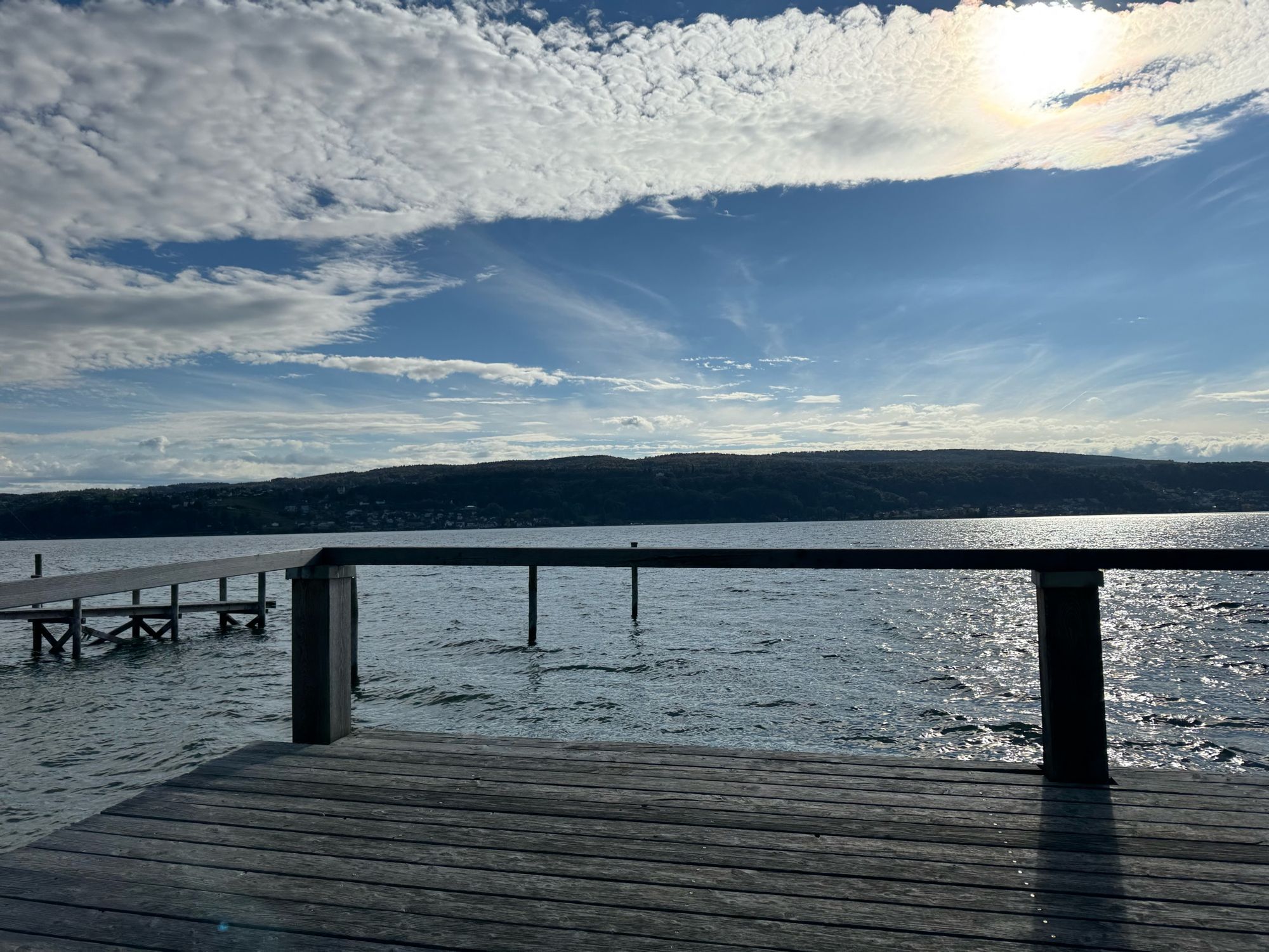 Das Bild zeigt einen hölzernen Steg, der in ein ruhiges Gewässer (den Bodensee) hineinragt. Der Himmel ist größtenteils bewölkt, mit einigen klaren Abschnitten, durch die die Sonne scheint. Im Hintergrund ist eine hügelige Landschaft zu sehen, die das Ufer des Wassers begrenzt.
