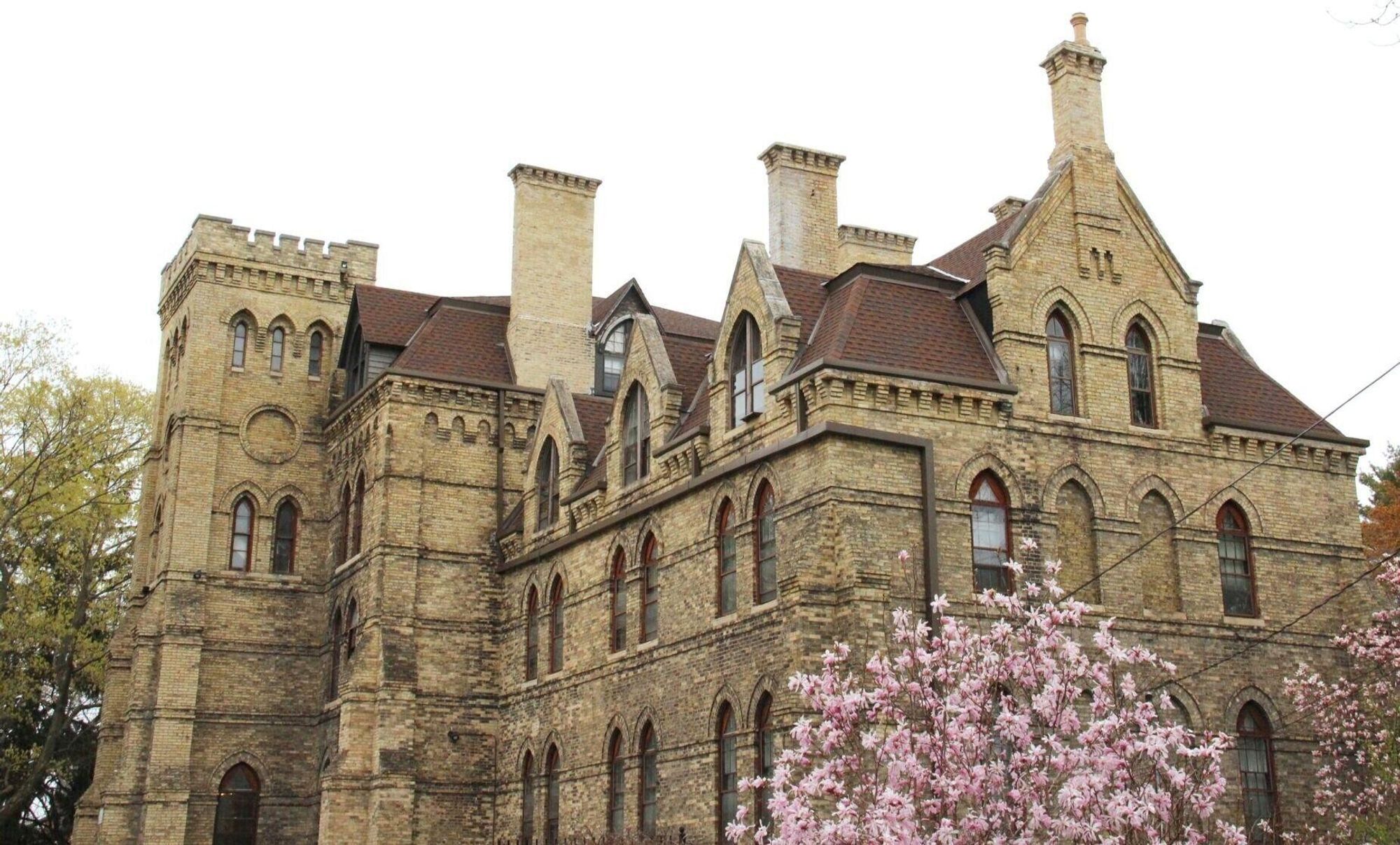 Newspaper photo showing a cream-brick, four-story building. It has a red roof, tall chimneys, ornate gables and lancet windows, in the Victorian Gothic Revivial style. A crenellated entrance tower is at left.