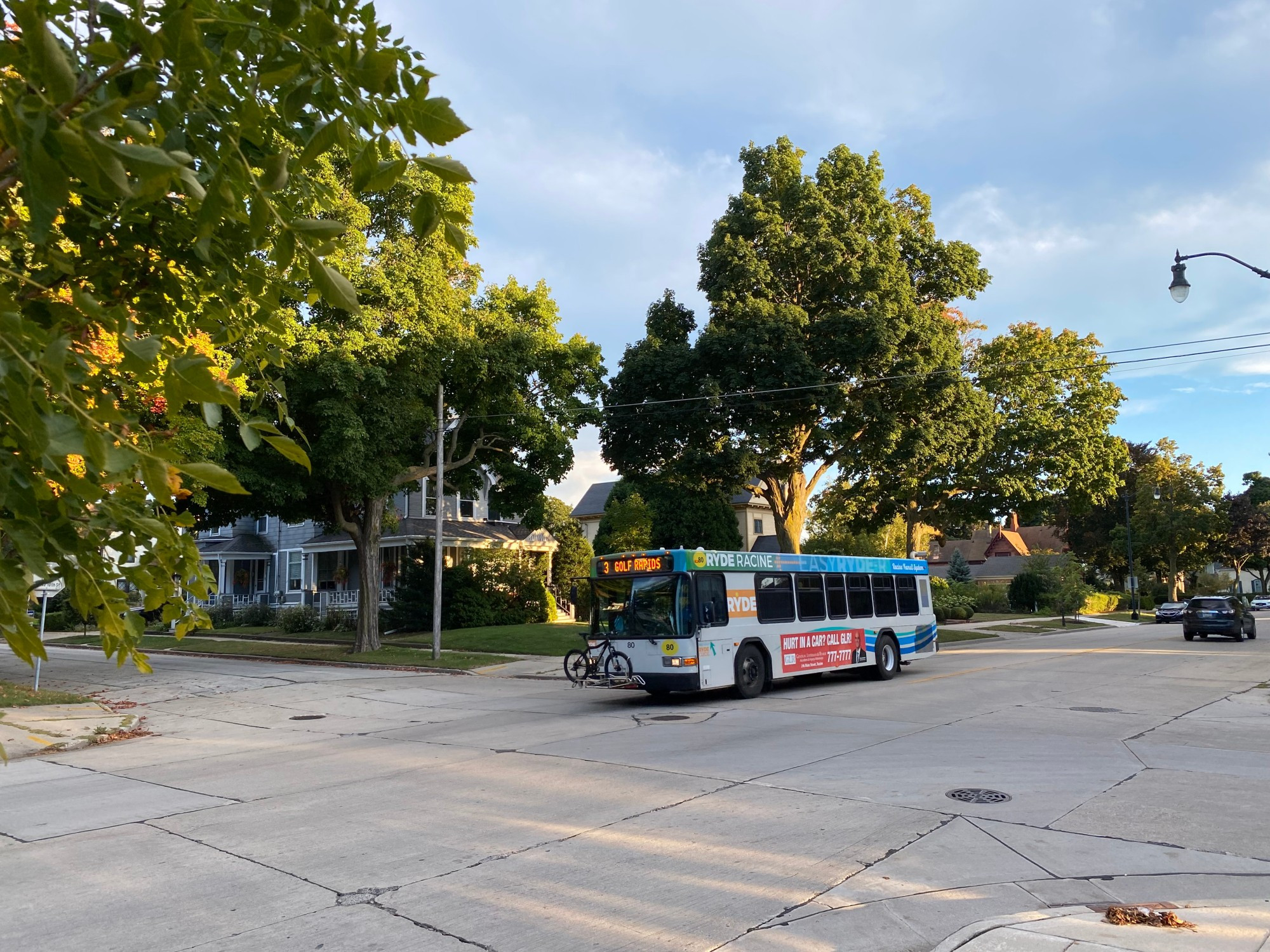 A local transit bus crosses through a street intersection in a residential area. A bicycle is mounted on the front of the bus. The destination board says "3 Golf Rapids". Trees in the foreground have a hint of fall color to them and obscure a row of Victorian houses behind the bus.