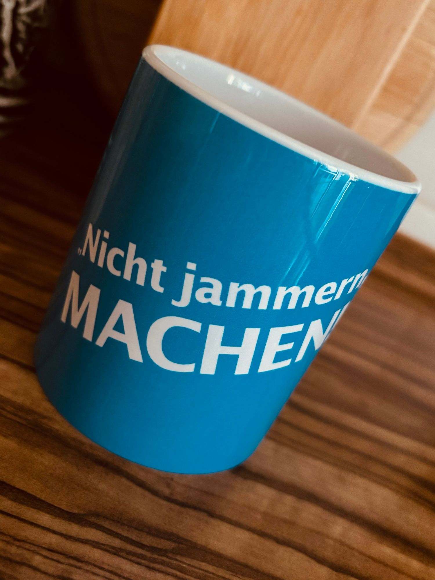 Ein blauer Becher mit dem Text "Nicht jammern, MACHEN" aufgedruckt, auf einer Holzoberfläche platziert.