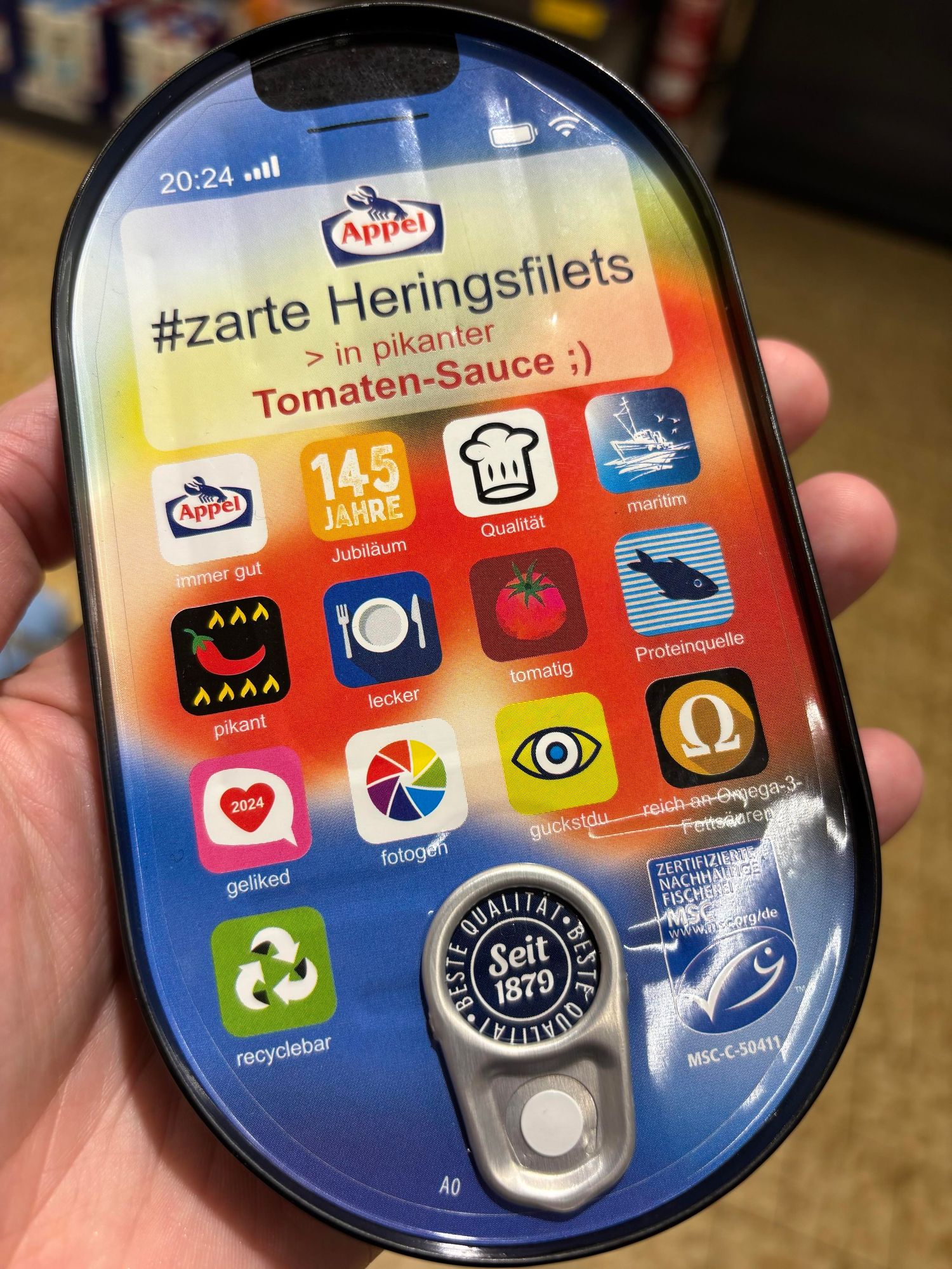 Eine Hand hält eine Dose Heringsfilets der Marke Appel in würziger Tomatensauce. Die Dose verfügt über ein farbenfrohes Design, das einem Smartphone-Bildschirm ähnelt, mit verschiedenen Symbolen, die Qualität, Zutaten und Nachhaltigkeit darstellen. Es hebt das 145-jährige Jubiläum der Marke hervor. 