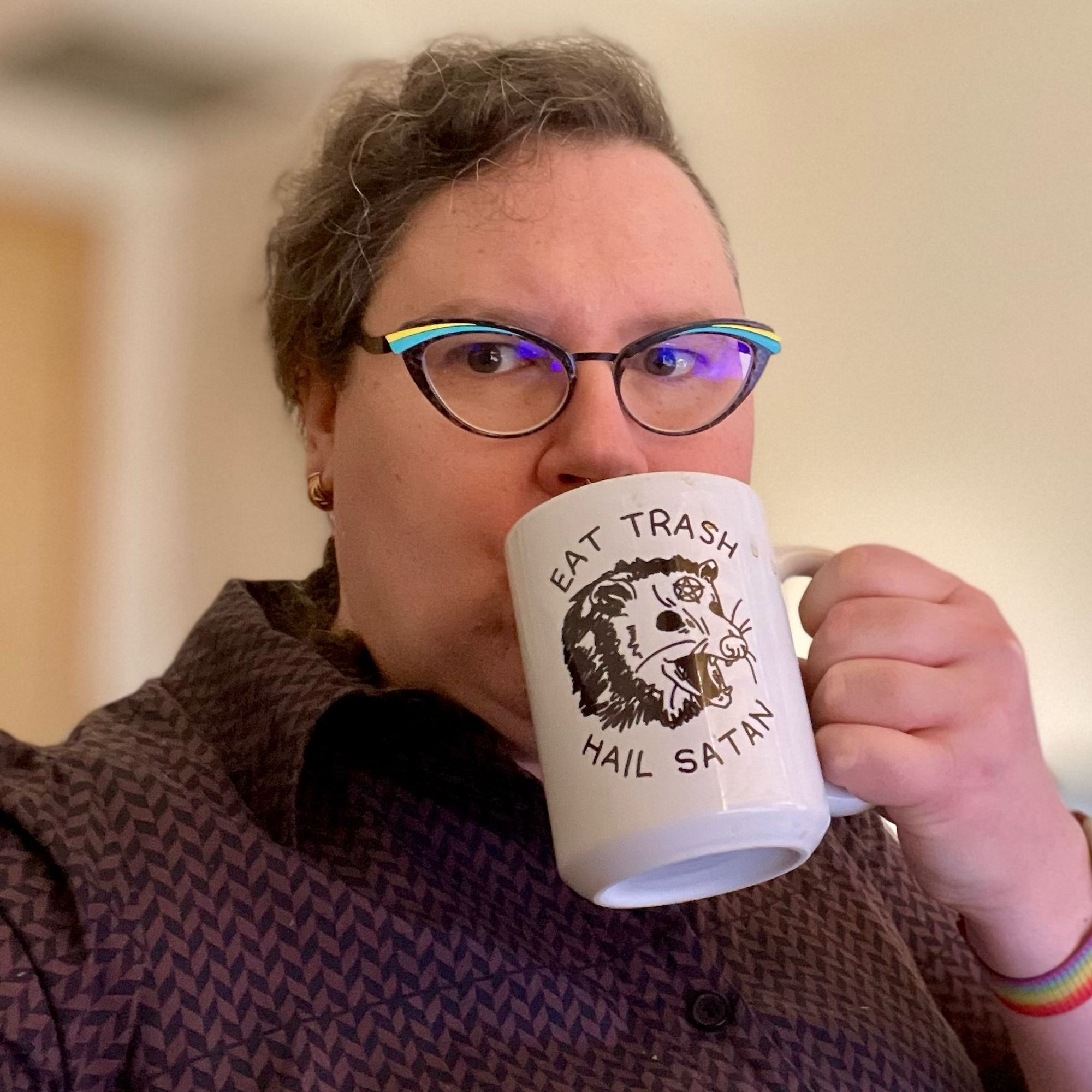 A middle-aged white woman with braided curly hair, cat-eye frames, and a brown checked shirtdress drinks coffee from a mug with a line drawing of a screaming opossum with a pentagram on its forehead, captioned “EAT TRASH / HAIL SATAN”