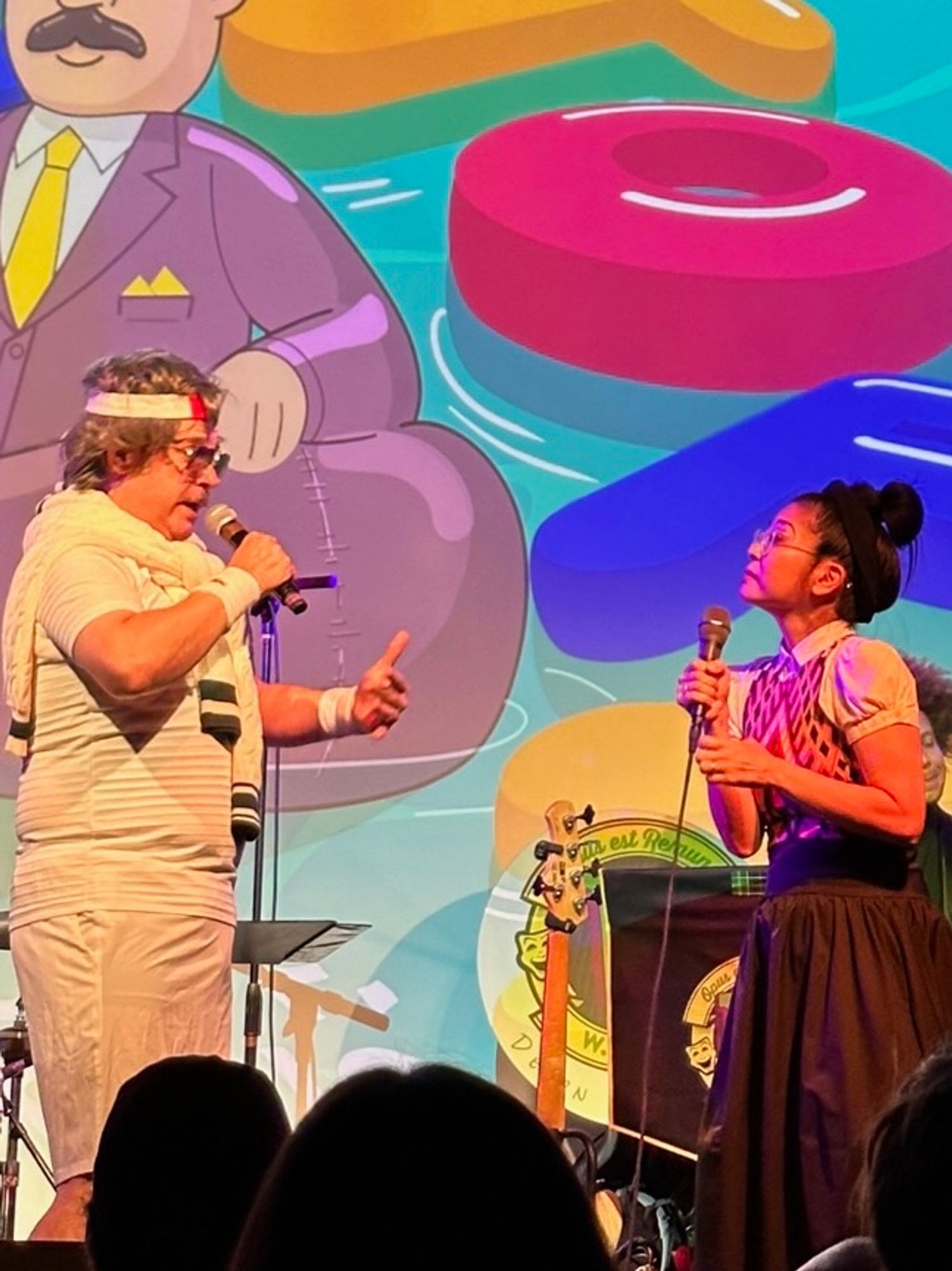 Two performers with mics onstage in front of a colorful projection.