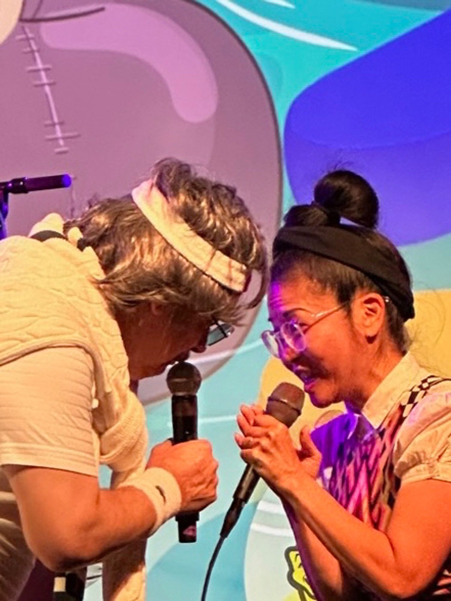 Two performers with mics standing close together whispering into them.