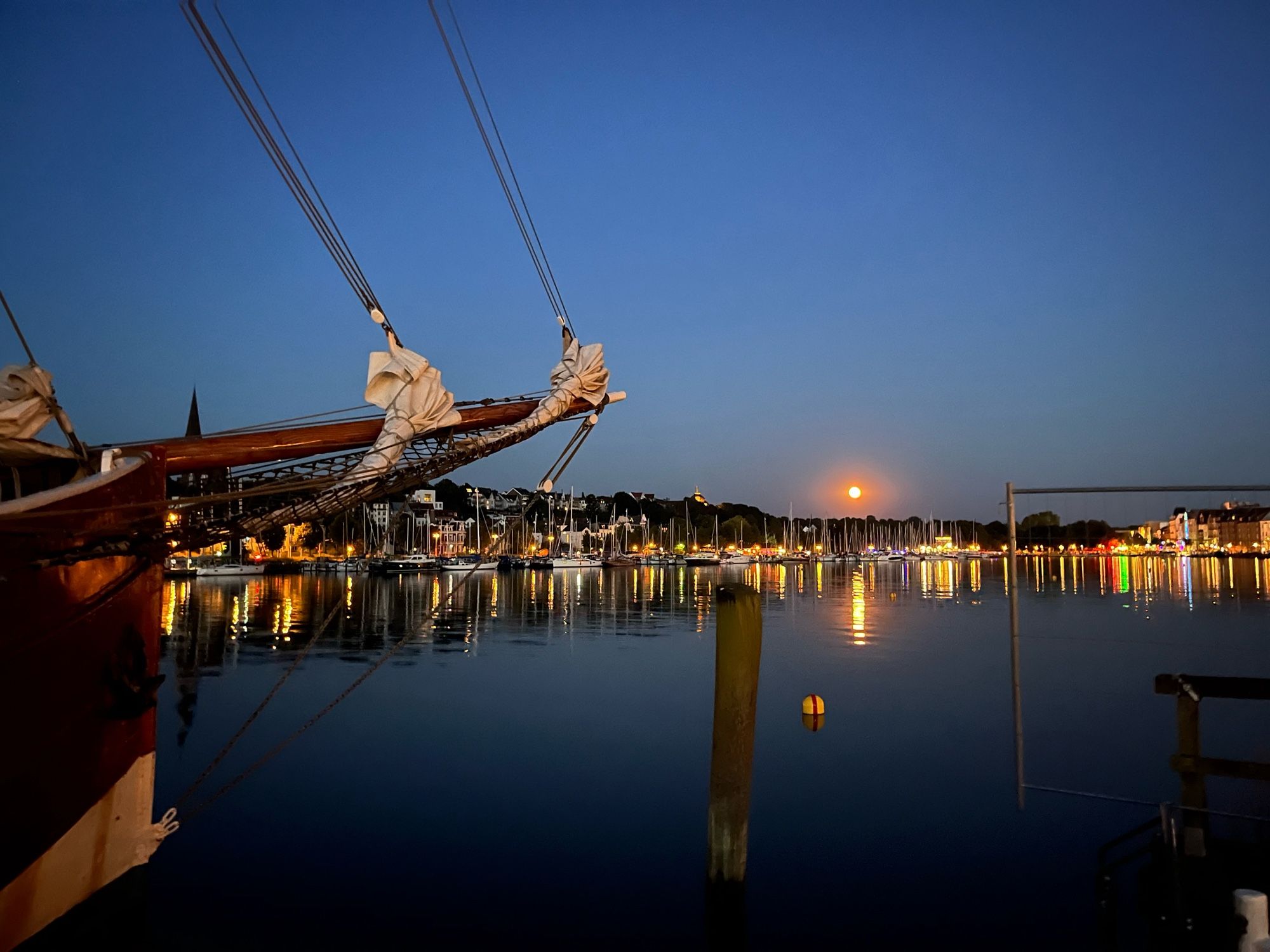 Blick über den Flensburger Hafen bei Nacht, im Vordergrund der Bug eines Oldtimer Seglers, im Hintergrund spiegeln sich Mond und die Lichter der Stadt in der Förde.