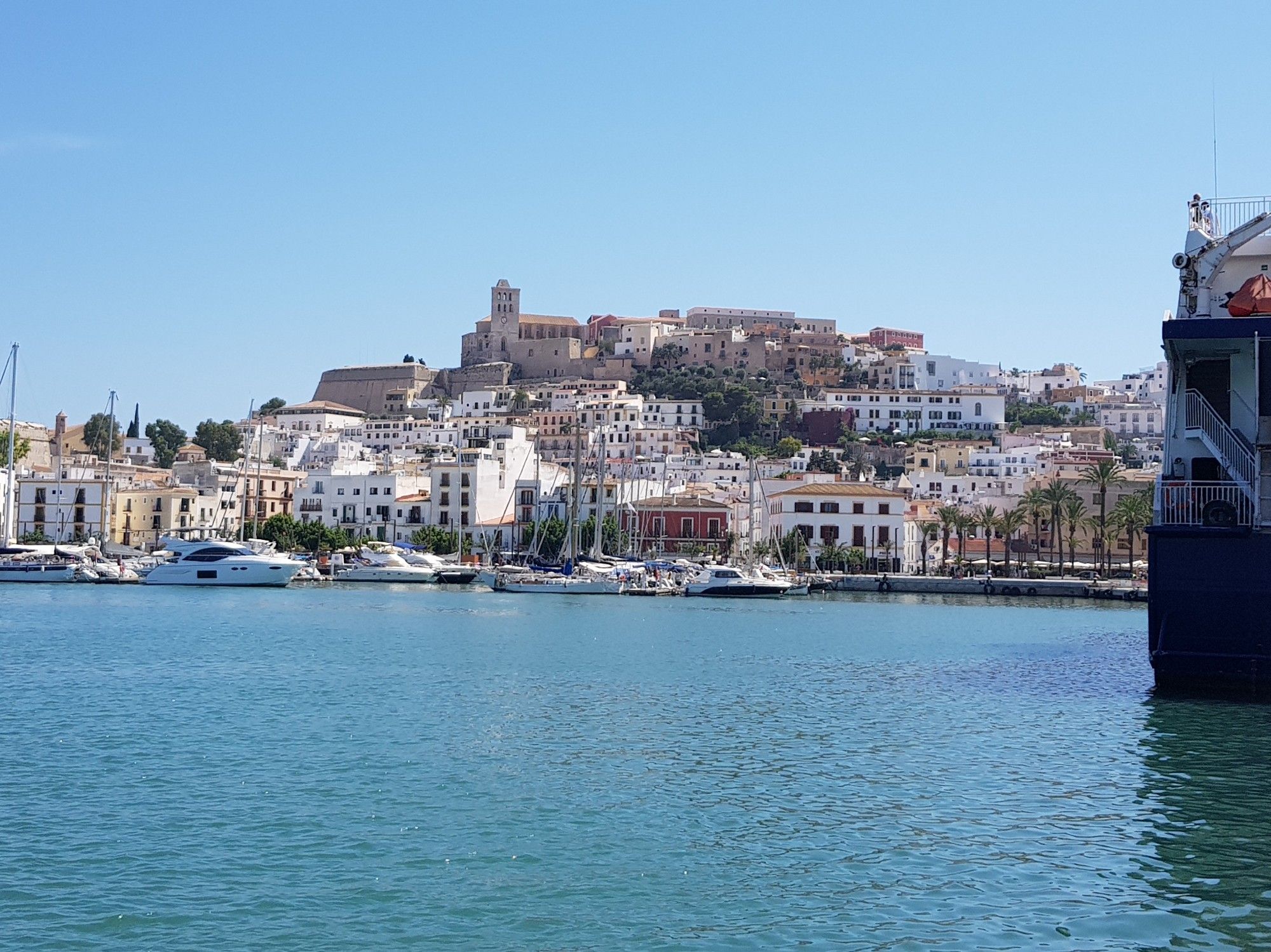 Blick auf den Hafen von Ibiza Stadt. Einige weiße Jachten liegen vor Anker. Dahinter meist weiße Häuser. Darüber eine Burg. Strahlend blauer Himmel und blaues Meer.