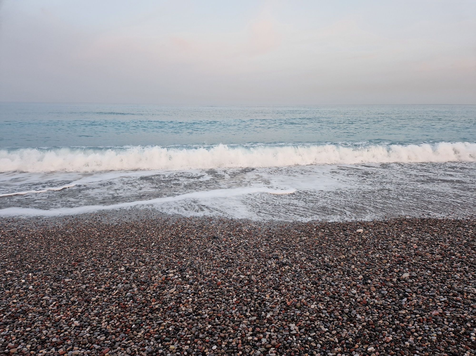 Stürmischer Kiesstrand auf Rhodos. Eine recht große Welle bricht gerade. Es ist stark bewölkt und sehr windig.