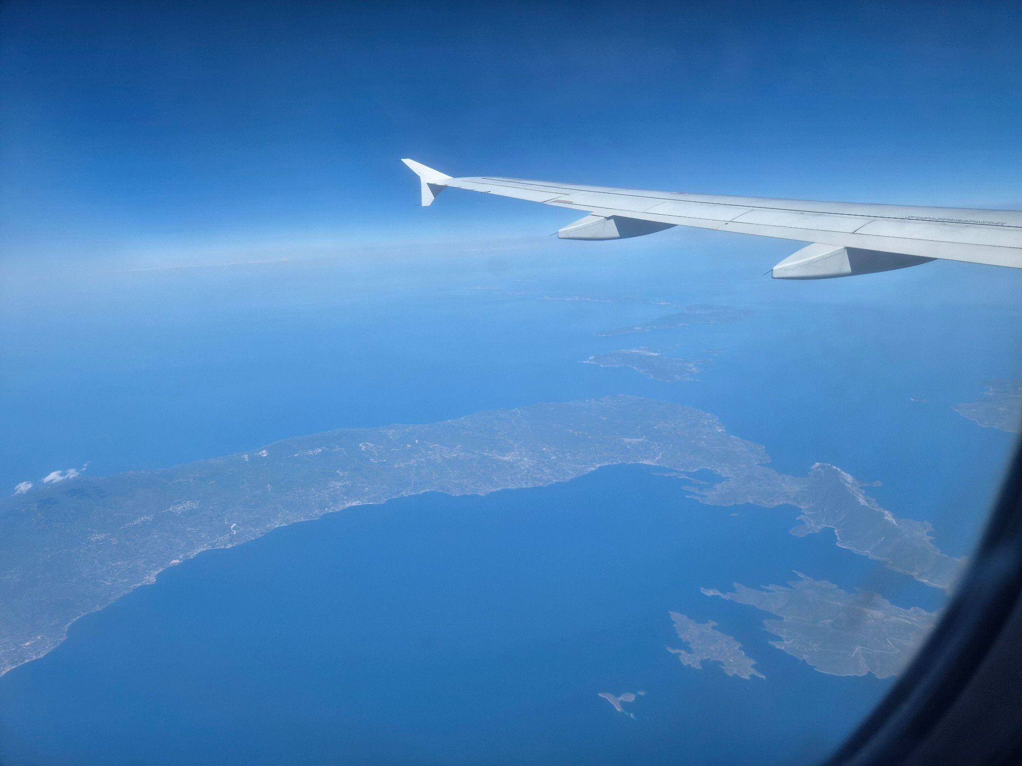 Aus dem Flugzeug fotografiert: linker Flügel eines Airbus, darunter eine Insel in der Ägäis. Blaues Meer und strahlend blauer Himmel. Am Horizont weiße Wolken.