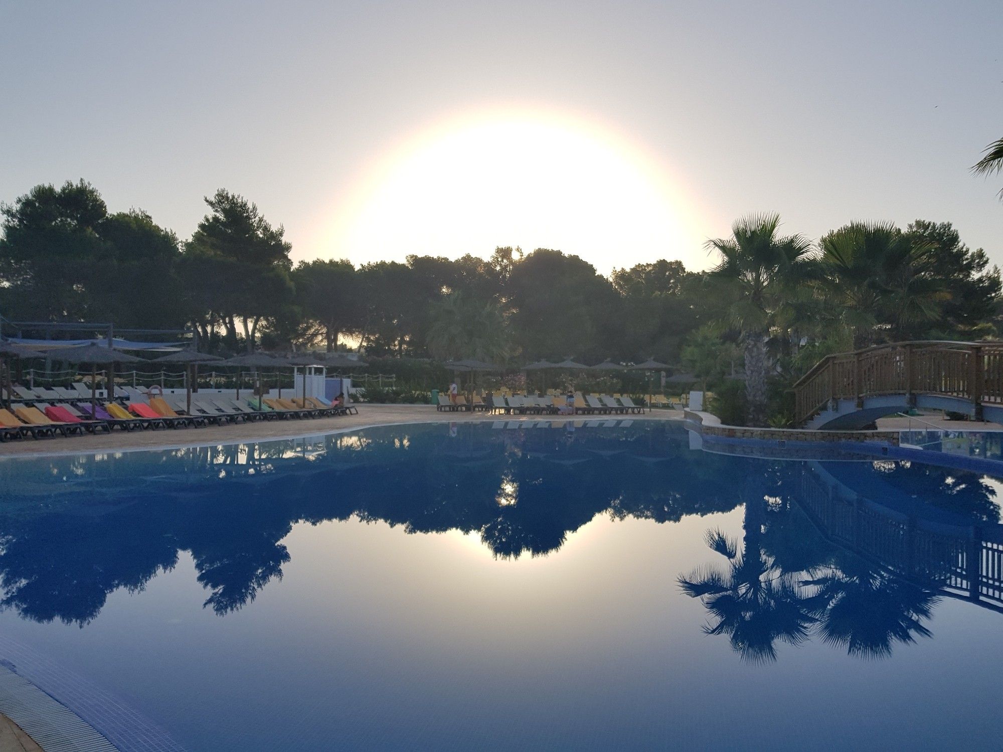 Sonnenaufgang über dem Hotelpool. Die Palmen spiegeln sich im Wasser. Auf einigen Liegen sind bereits Handtücher. Kein Mensch zu sehen.