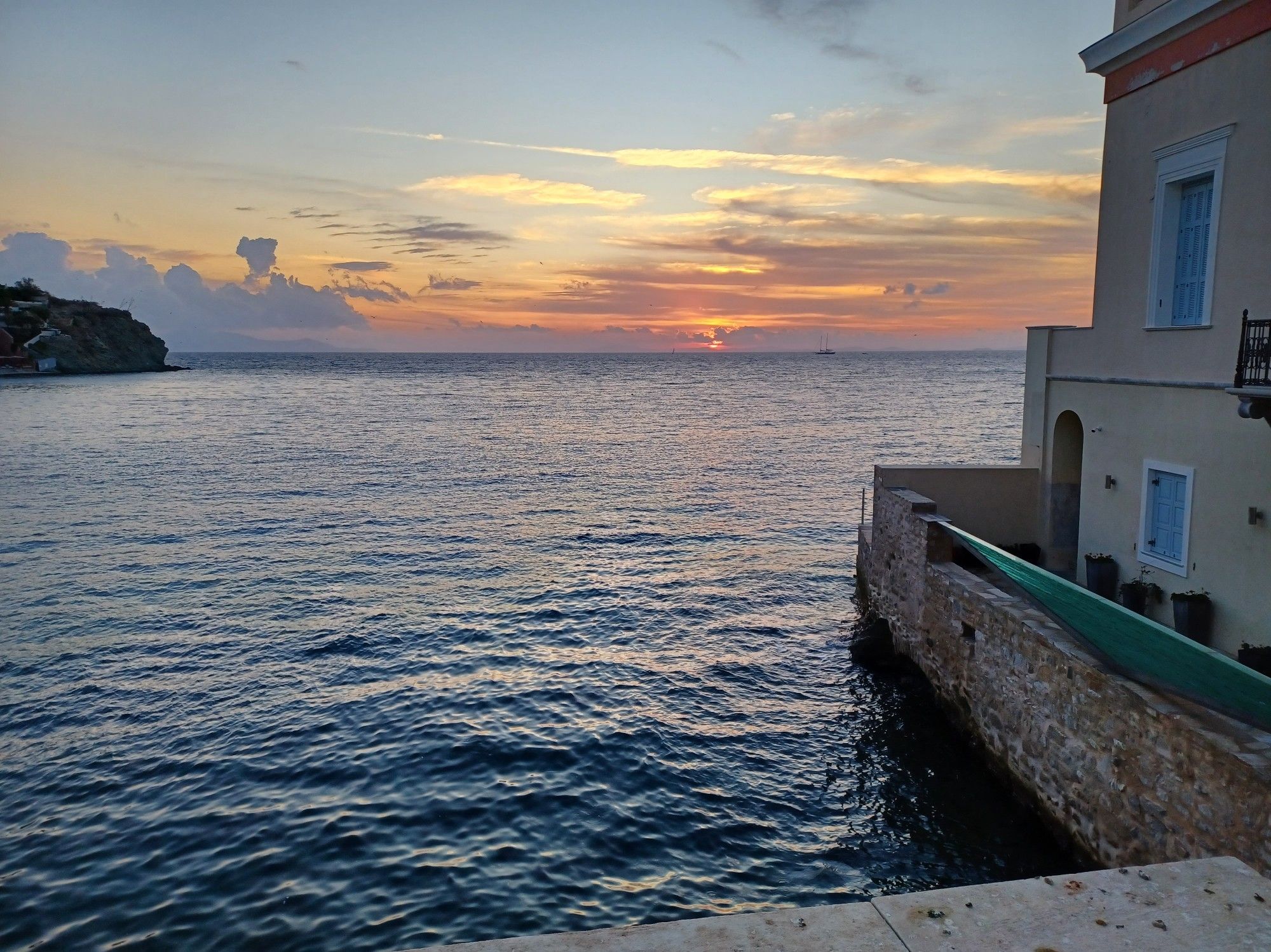 The sun having just risen behind clouds past a distant island. The point of view is from some coastal buildings. There is a distant sailboat passing by.