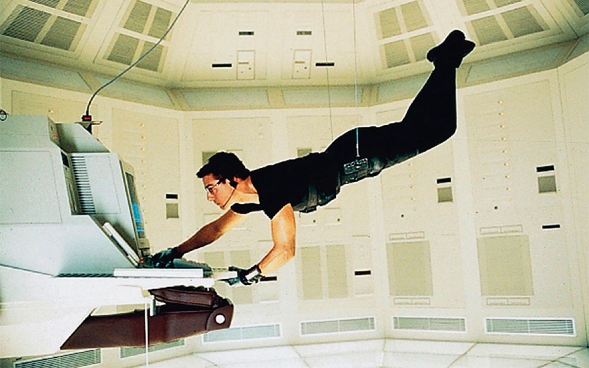 Tom Cruise in the first Mission Impossible movie on a wire stealthily using a computer keyboard. He's wearing all black outfit in an all white and cream room.