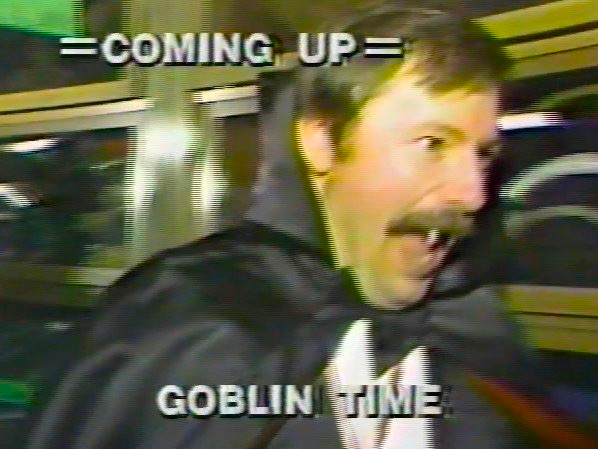 a man from the 80s in a vampire costume complete with fake fangs under a real stache. text overtop reads in all-caps: COMING UP GOBLIN TIME