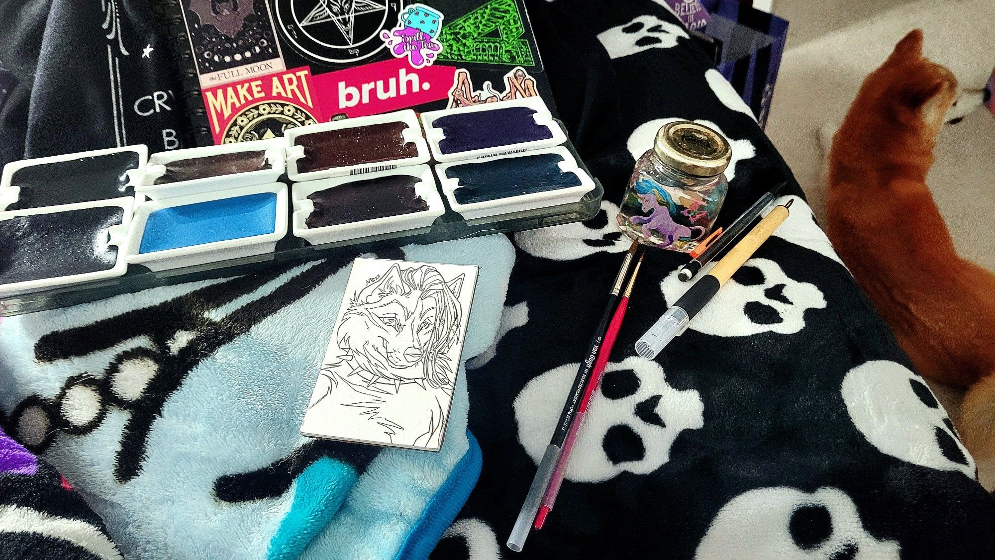 Casual photo of the artist's work space which appears to be a bed covered in colorful fuzzy blankets. Piece being worked on is a small unfinished drawing of a cartoon dog in ink work. There are artist's materials arranged around the piece including 2 small round brushes, a fine Japanese brush pen, mechanical pencil, a small jam jar repurposed for holding water ( lid is on and jar decorated with unicorn stickers ), a plastic tray holding a spread of large watercolor pans in the colors the artist intends to use, and a peek at the artist's personal sketchbook covered in ephemeral stickers. On the upper right of the photo on the floor can be seen the artist's pet shiba inu dog laying on the carpet. She is very orange and smiley.