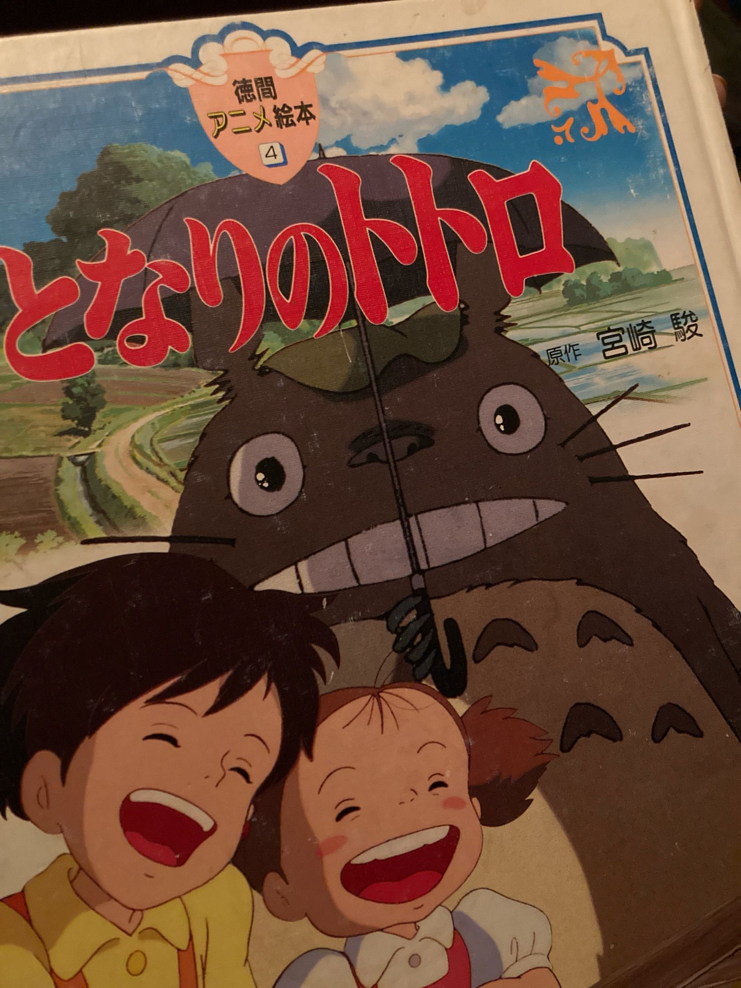 A photograph of the slightly worn front cover of a Japanese "My Neighbor Totoro" picture book. Besides the title, publisher, and creator's name, it shows Satsuki, Mei, and of course the large Totoro, who is holiding an umbrella, against a bright countryside.