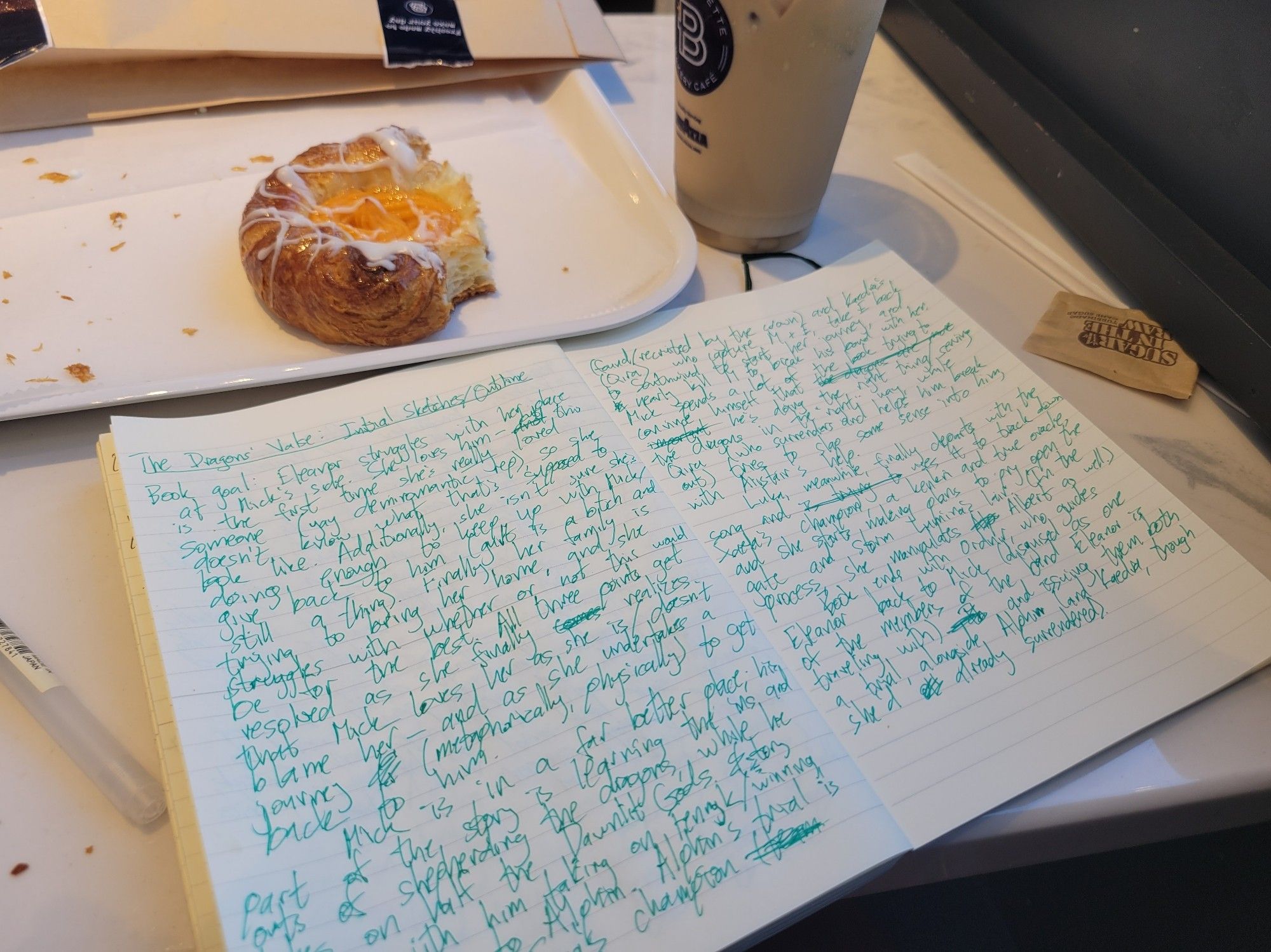 A notebook sits open on a messy table. Its pages are covered with writing in green ink; a pen sits to the left of the book. Above the book, also on the table, are a large iced latte in a plastic cup and a white tray holding danish with an orange center.