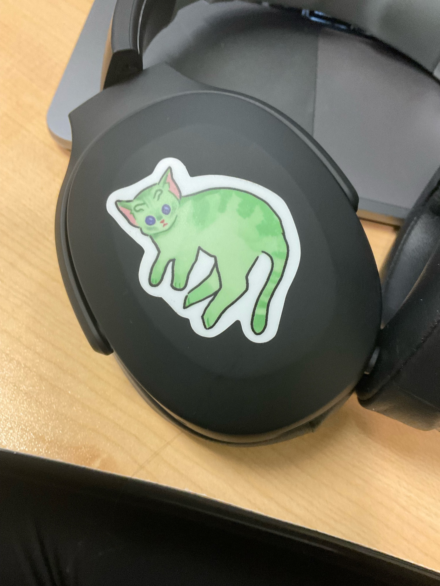 A sticker of a digitally illustrated alien cat on black headphones. The cat is green and laying on its side. 