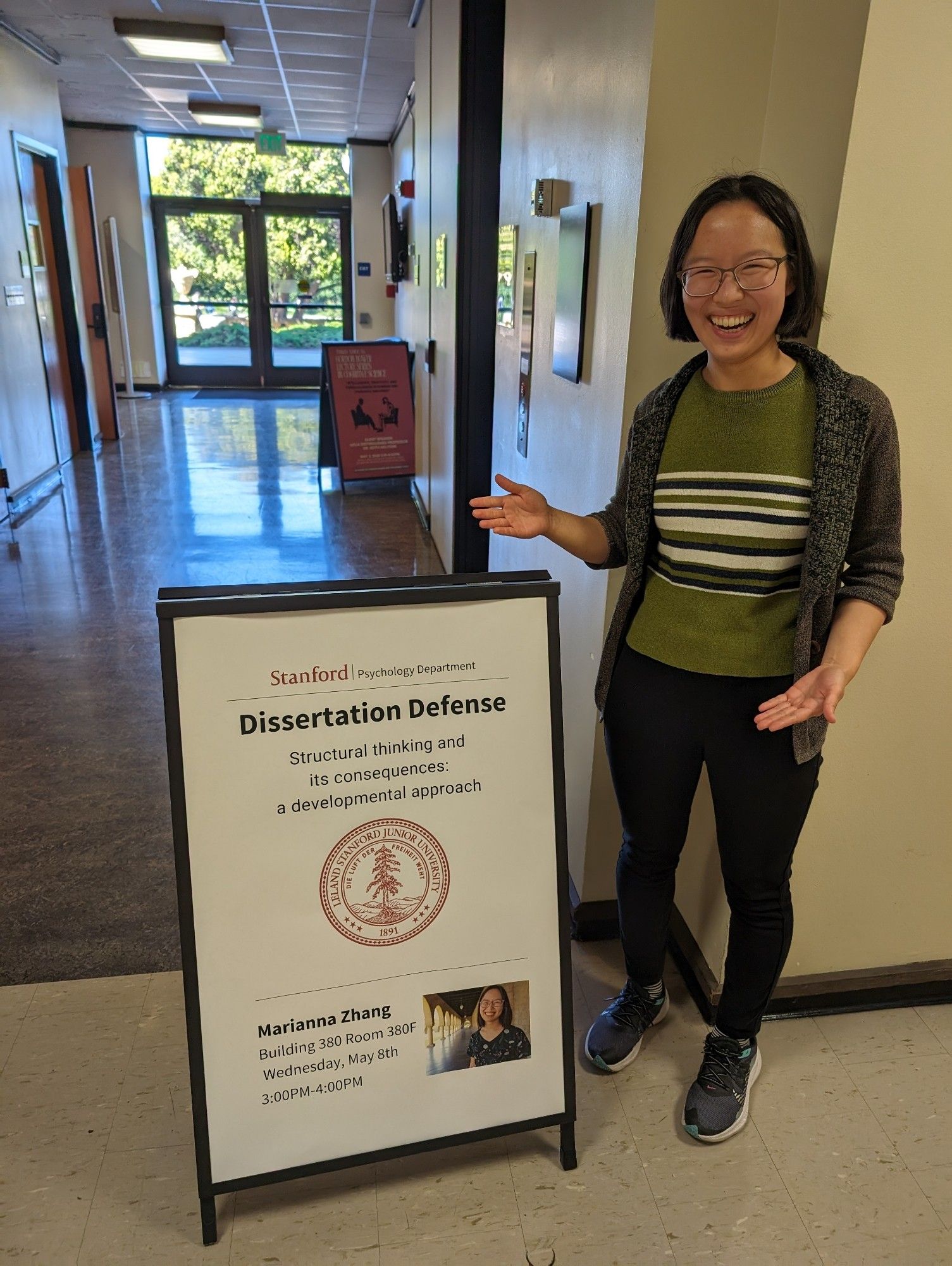 asian woman grinning while gesturing to poster stand reading dissertation defense