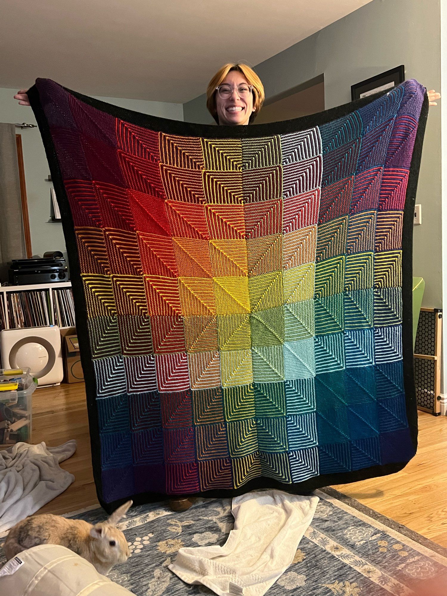 Gale holding a knitted rainbow blanket in front of them. It’s like five feet tall. A house rabbit walks along the rug in front of Gale, refusing to acknowledge their tremendous achievement.