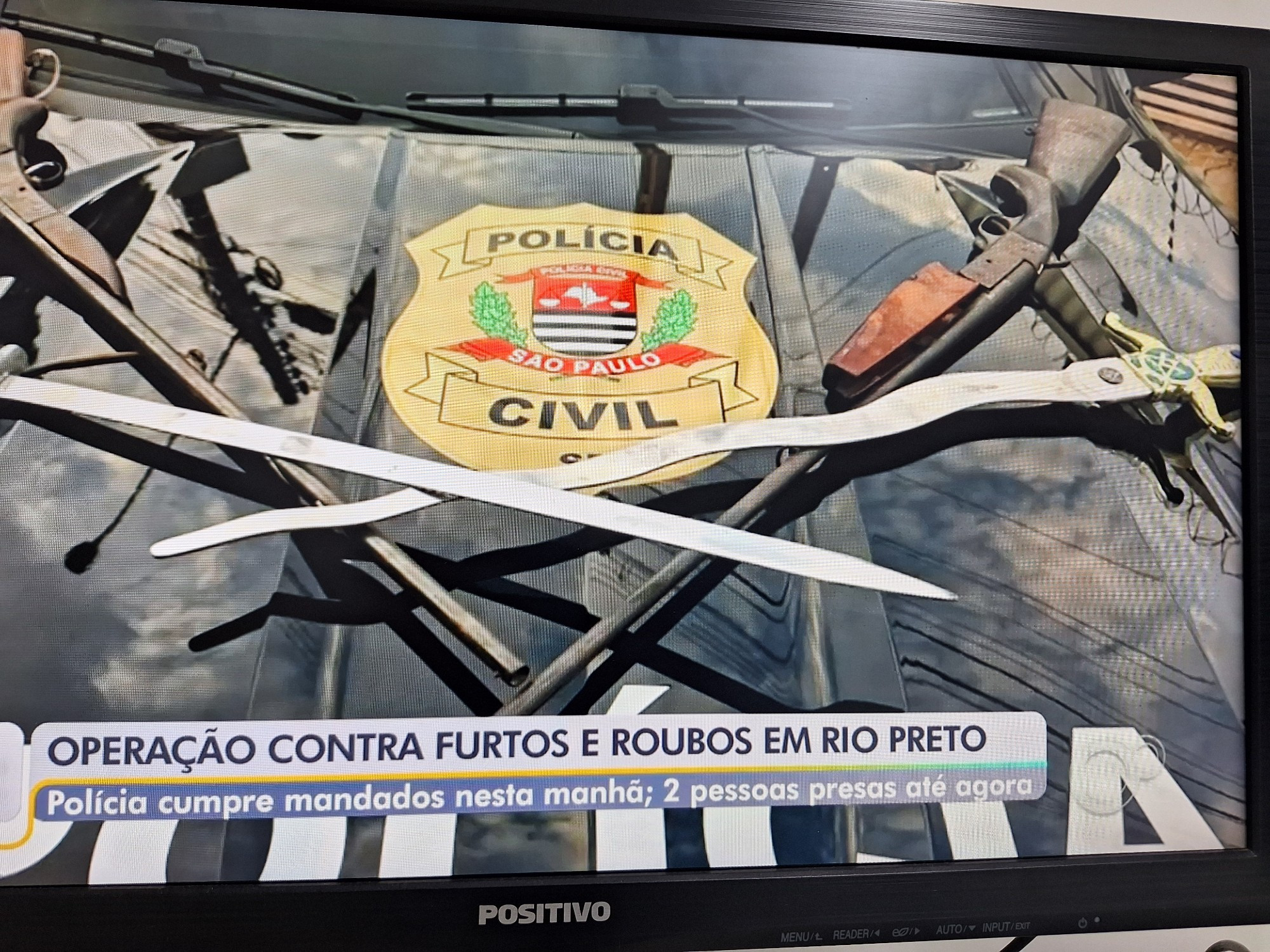 Foto de uma reportagem na TV. A imagem mostra o capô de um carro preto da polícia civil, com um logotipo dourado da Polícia Civil. Em cima desse logotipo há duas armas de fogo tipo espingardas cruzadas e duas espadas. Uma delas é ondulada, prata, com a empunhadura dourada e um logotipo da maçonaria .