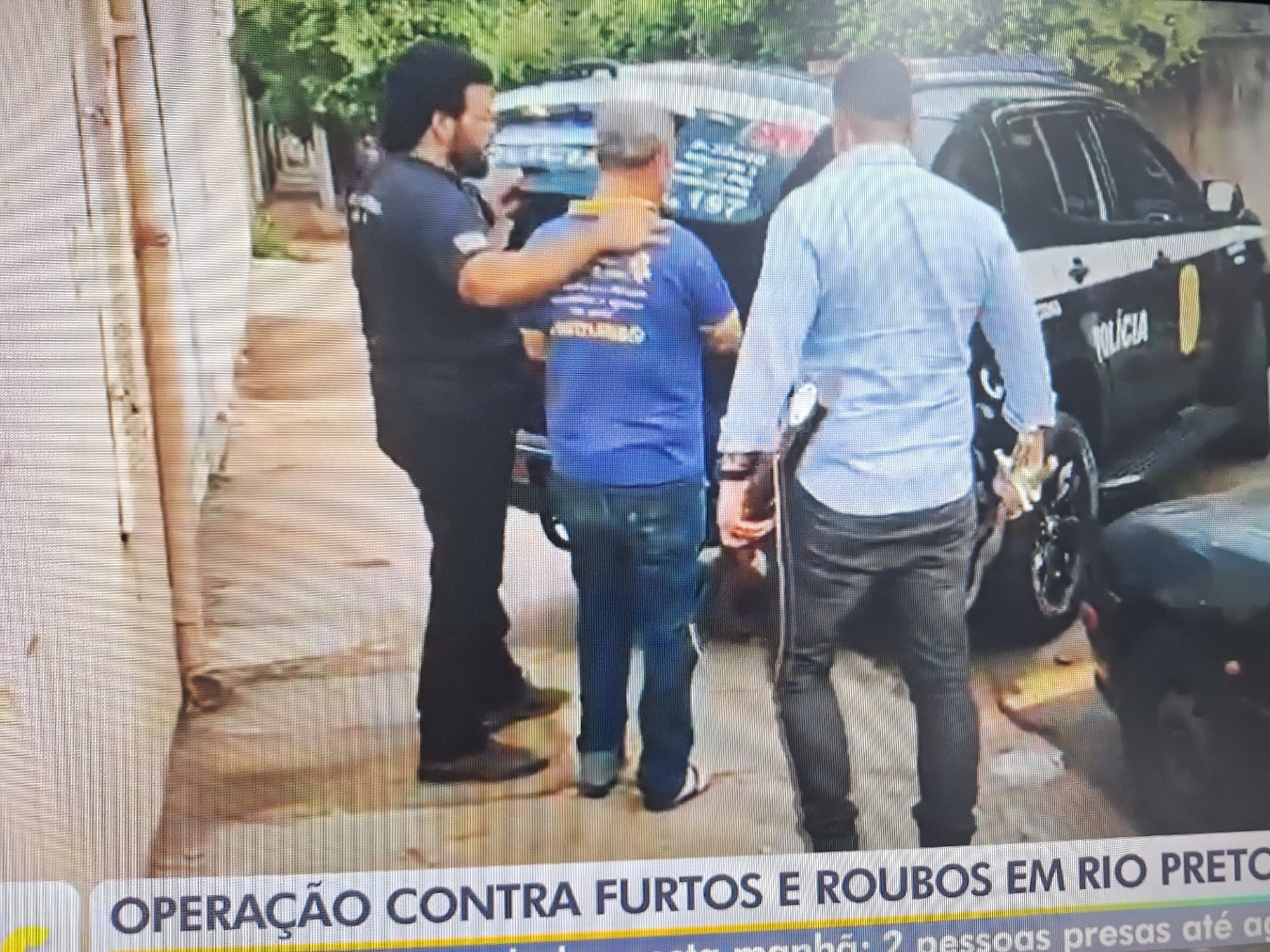 Imagem da reportagem na tv. Na imagem já um carro de polícia preto, com o porta-malas aberto. Um policial de camiseta preta, de costas, está com as mãos no ombro de um homem de azul e boné. Ao lado, um outro homem que deve ser policial, também de costas, está com uma arma de fogo na mão esquerda e duas espadas na mão direita. Ele veste uma camisa social azul clara e calça jeans.