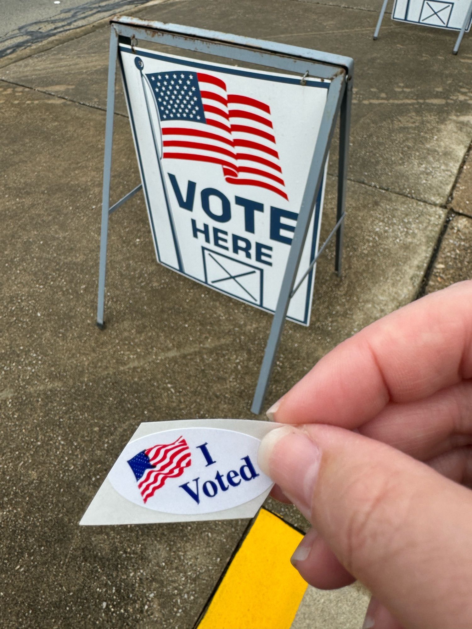 Picture of a "Vote Here" sign with a hand in front of it holding a "I Voted" sticker