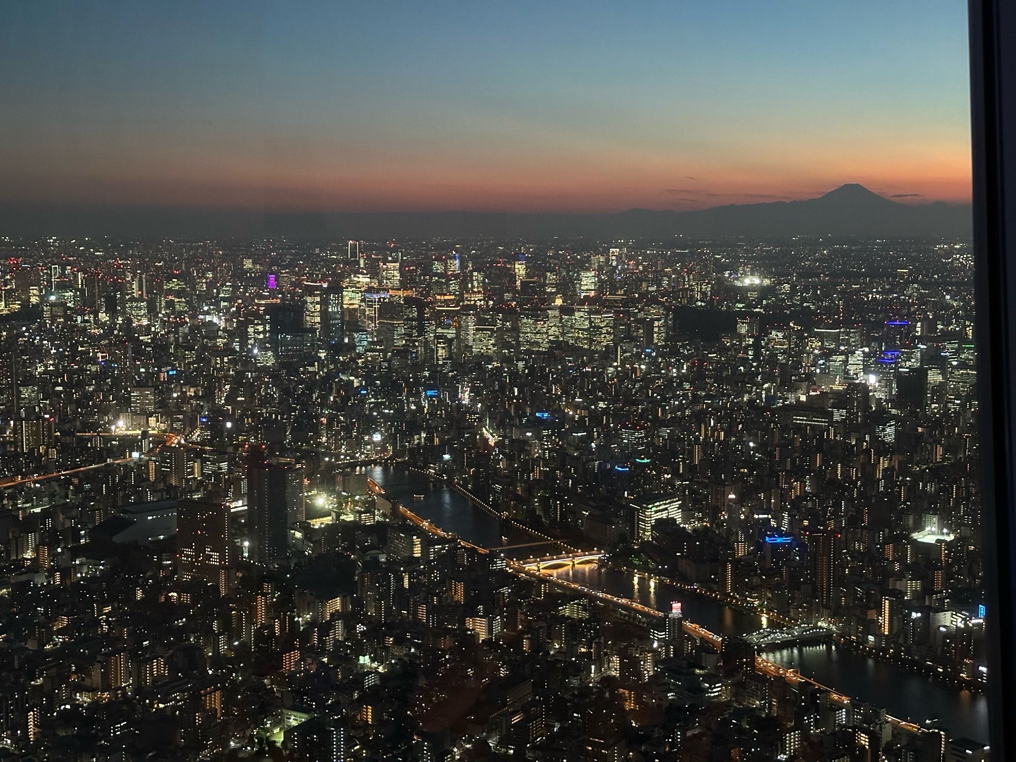 A view of Tokyo at sunset, taken from the observation deck at Tokyo SkyTree, 451m up. Visible at centre-left is Tokyo Tower, illuminated purple; at upper right, Mount Fuji.