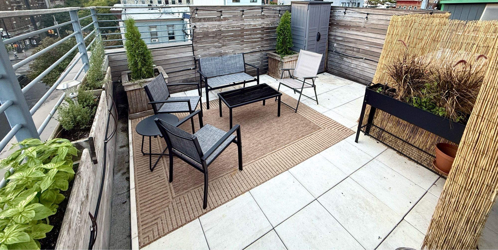 same corner of roof cleaned up with nice seating and a coffee table, rug, plants, and a reed wall hiding the big AC units