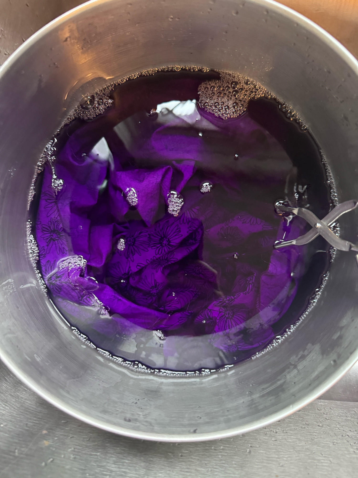 Cotton fabric dyed purple in a color fixative solution in a stainless steel pot. A pair of stainless steel tongs for stirring and retrieving the fabric are also in the pot.