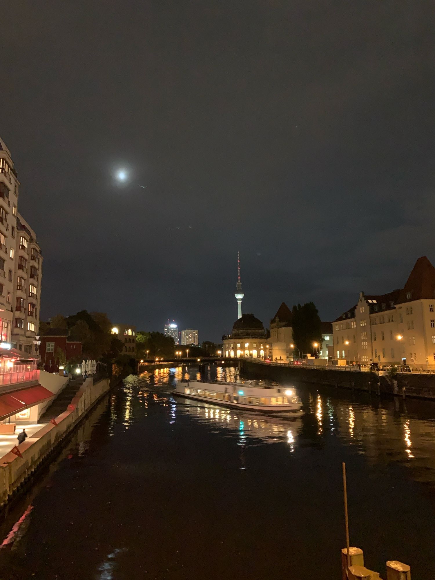 Foto von Berlin bei Nacht. Die Spree, mit einer Barkasse. Im Hintergrund der Fernsehturm und oben der Mond. An den Seiten links und rechts Gebäudesilouetten.