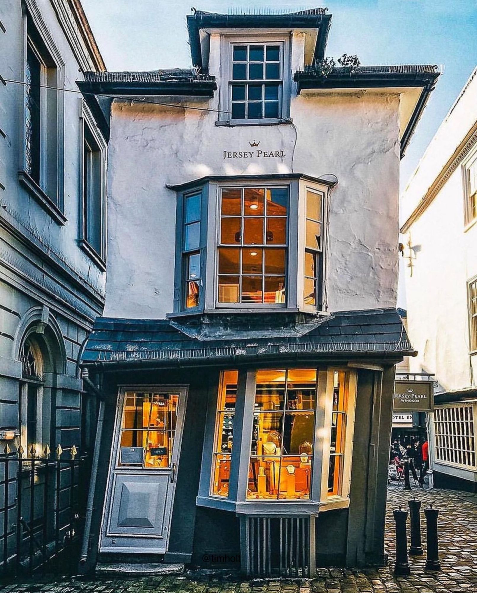 Picture shows an old small, white tall house leaning in a wonky fashion! Warm light glows from the windows… perfect place for a cuppa!  