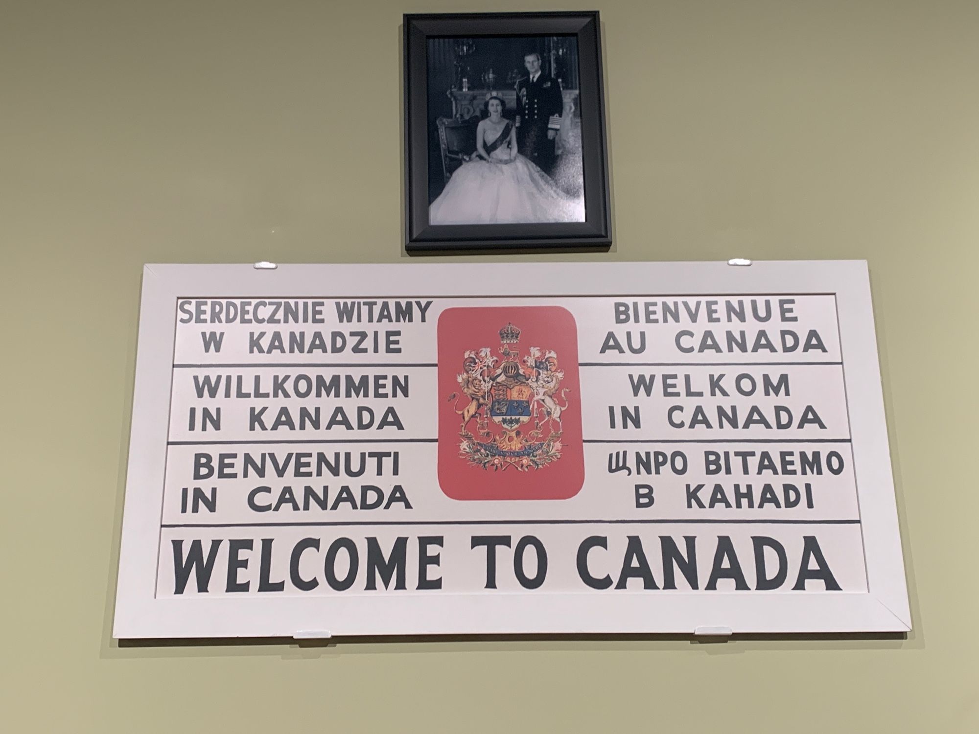 A replica of the Welcome to Canada sign from 1946 at Pier 21 in many languages with the typos kept as in the original. A photo of Queen Elizabeth II with Prince Philip is above, but she would not have been married yet, or have been Queen yet.